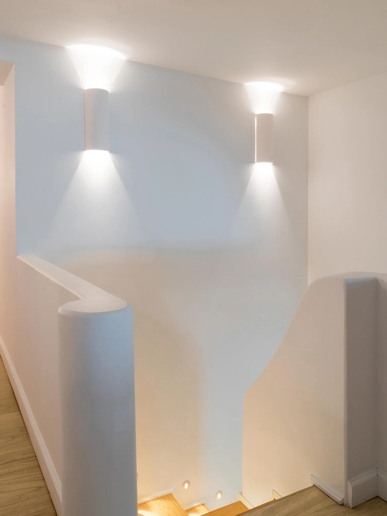 The upper level of the staircase featuring clean white walls and rhythmic up-and-down light patterns from modern cylindrical sconces. The image emphasizes the sophisticated lighting design and the quiet luxury of the space. Architectural photography 