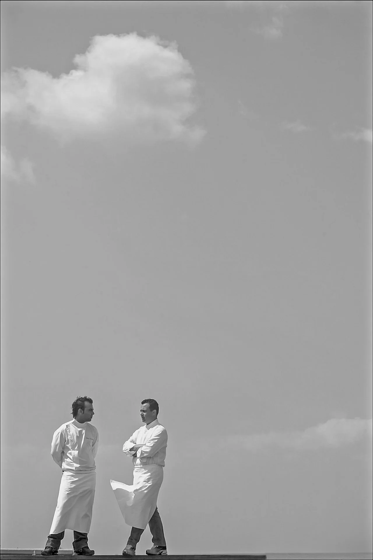 Minimalist editorial portrait of chefs Miltos Parikos and Giannis Karathanasos against a clear blue sky. A contemporary gastronomy photography study by Stavros Kostakis, emphasizing professional stature and the clean aesthetics of modern Greek culina