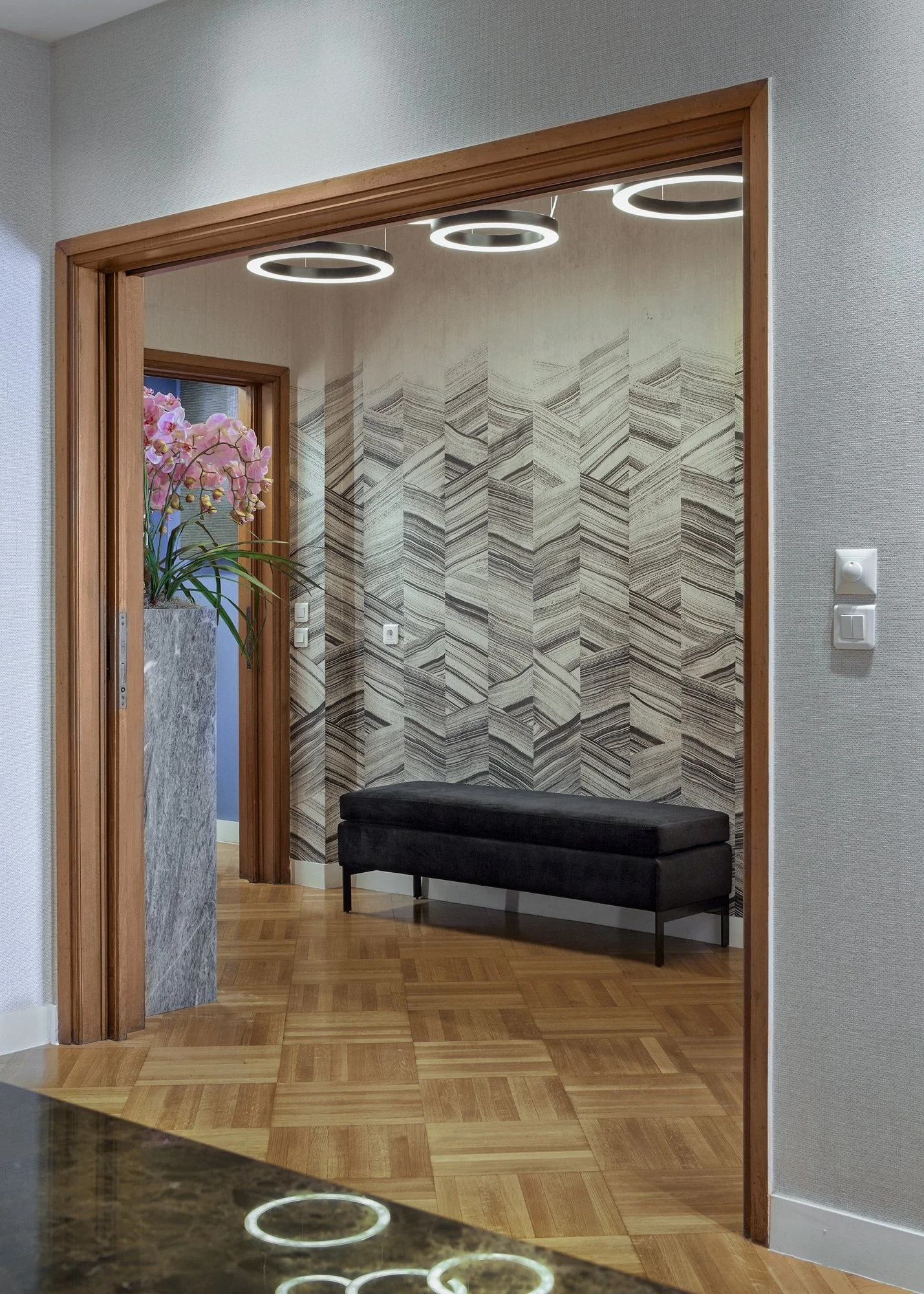 Professional architectural photography showing the perspective of a Kolonaki law firm. Features a dark velvet bench, signature chevron wallpaper, and circular ceiling lights, framed by a wooden doorway. Design by Sissy Feida.
