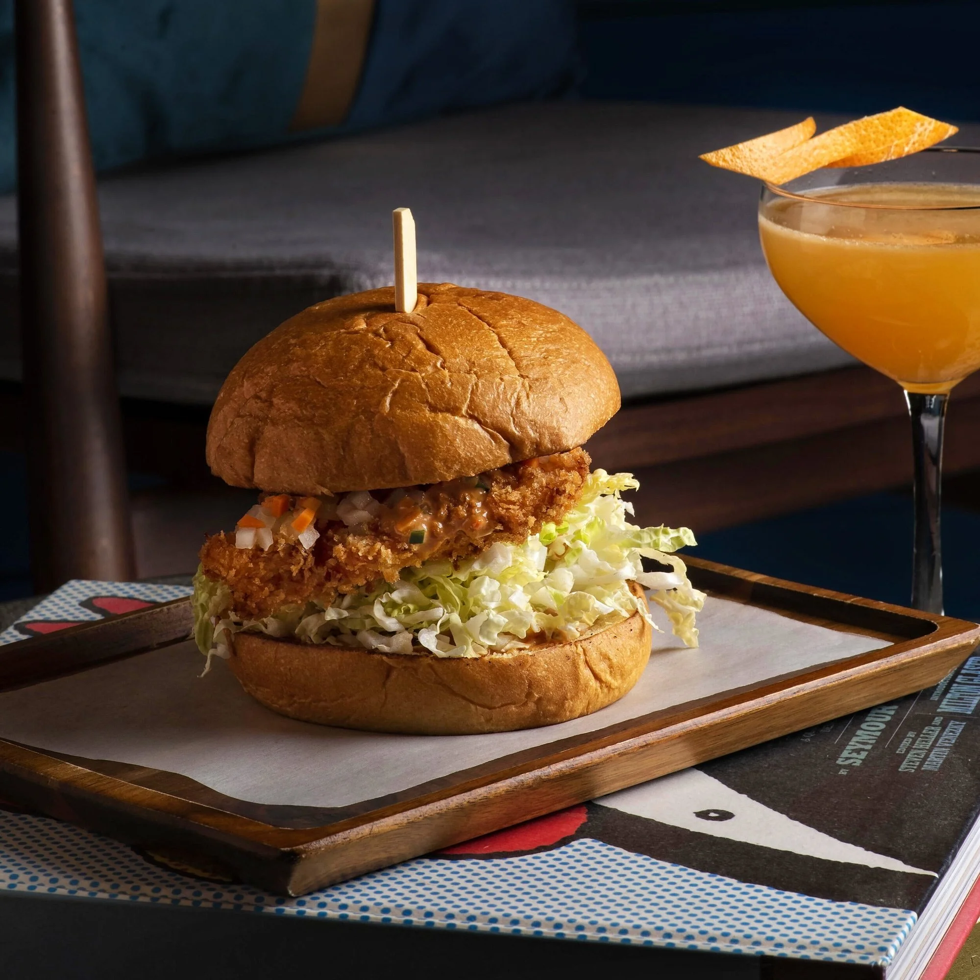 A gourmet crispy chicken burger with fresh slaw and pickles, served on a wooden tray alongside a sophisticated orange cocktail. Commercial photography for Royal Monkey by Stavros Kostakis.