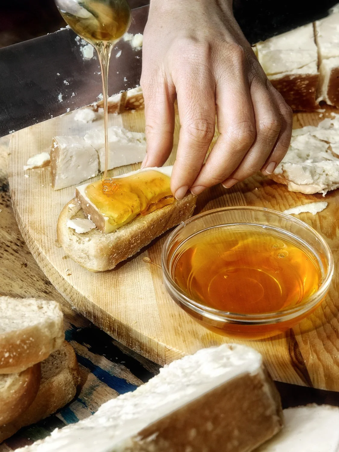Close-up of hands pouring honey over traditional Karavas rusks and cheese on a wooden board, shot by Stavros Kostakis.