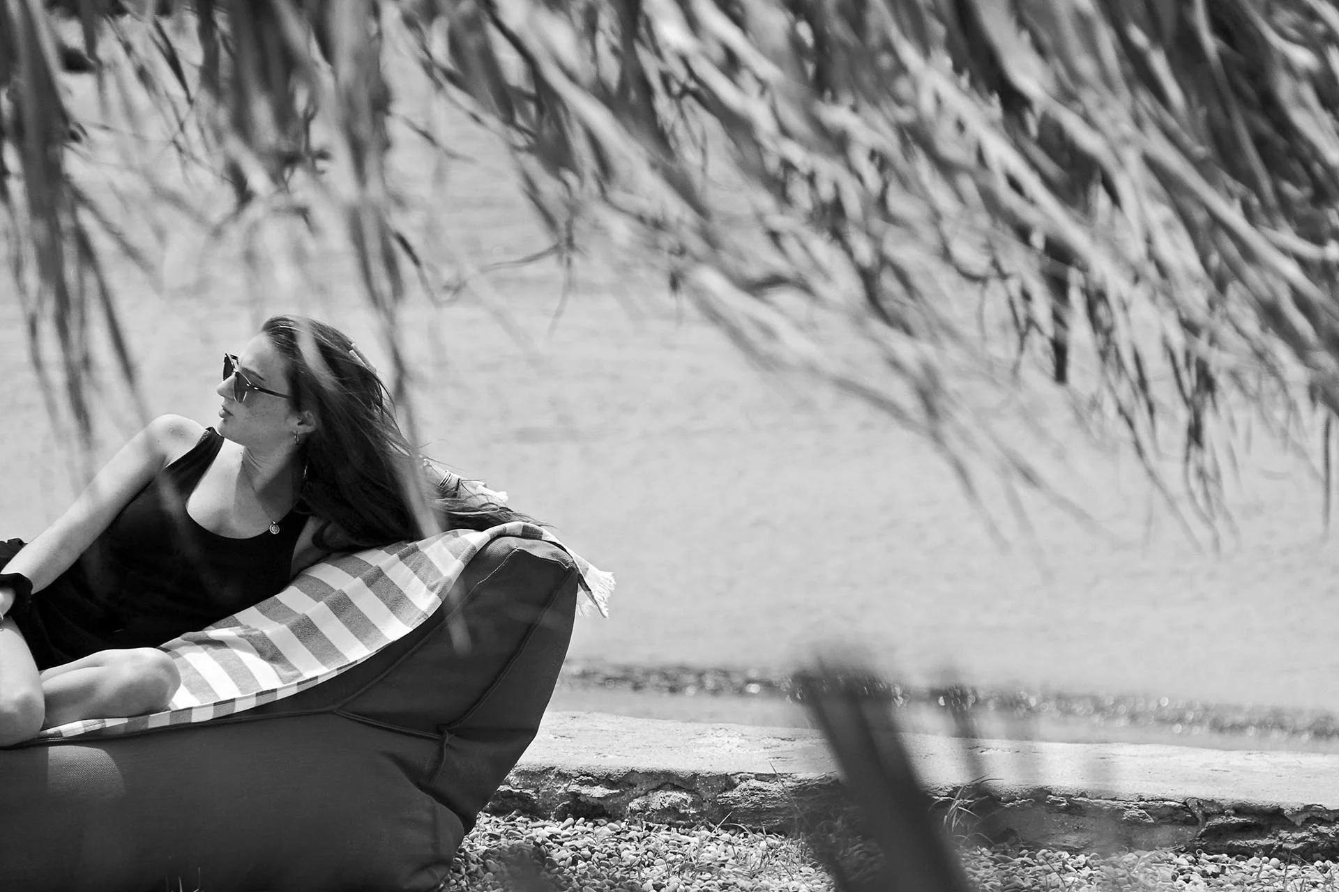 Candid black and white beach lifestyle photography at Bianco Beach House Tinos, featuring a woman relaxing on a sunbed by the Aegean sea, framed through natural textures to create a cinematic documentary feel of island leisure, by Stavros Kostakis.