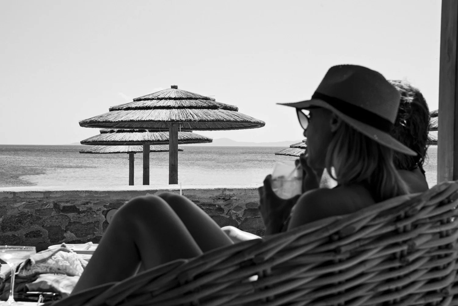 Sophisticated black-and-white recording of a woman's lifestyle with a hat, looking and drinking her cocktail at the Aegean Sea at Bianco Beach House, emphasizing a timeless, unpredictable moment of Cycladic relaxation, by Stavros Kostakis.