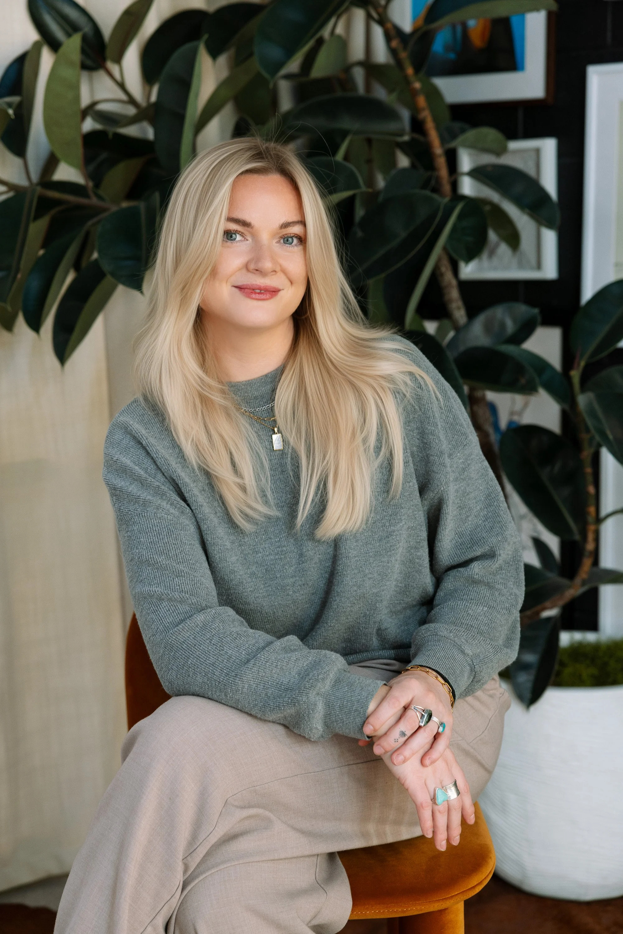 A woman with long blonde hair, wearing a gray sweater and beige pants sits on a brown chair in front of a large green leafy plant, with framed artwork on the wall behind her.