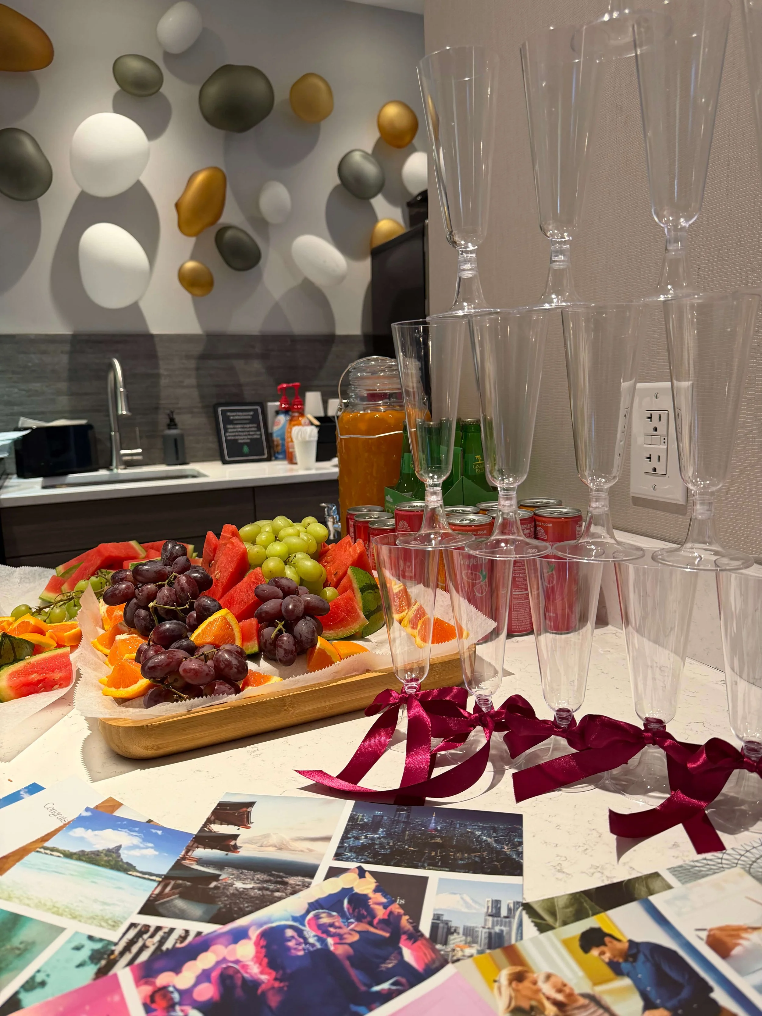 A table with an assortment of fresh fruits including grapes, watermelon, oranges, and other sliced fruits, alongside empty champagne glasses, postcards, and condiments, with a modern decorative wall featuring colorful abstract shapes in the backgroun