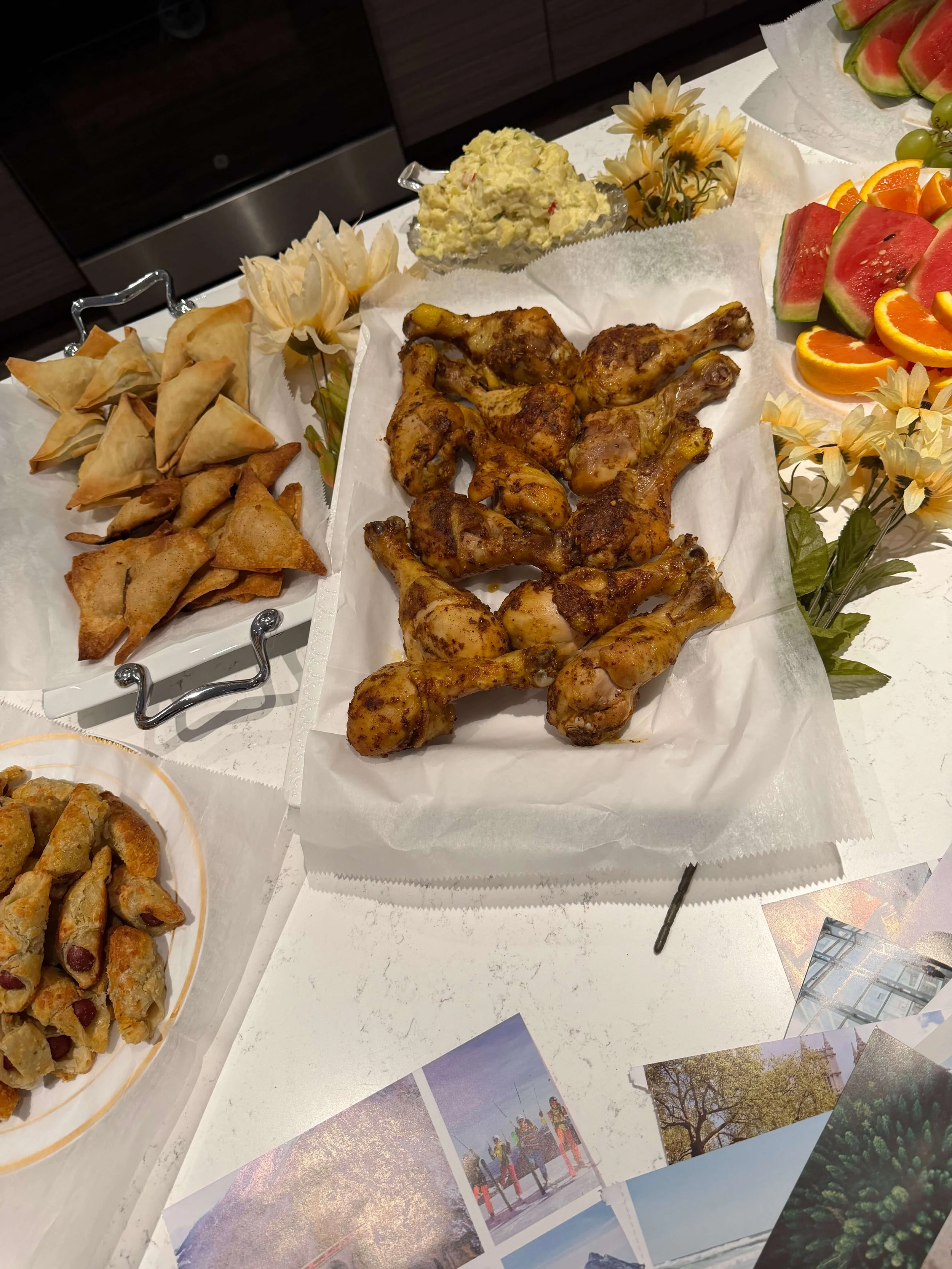 A table with various foods, including grilled chicken drumsticks, watermelon slices, orange and grape slices, potato chips, and a dish of potato salad, decorated with artificial flowers and several printed photographs of outdoor scenes.