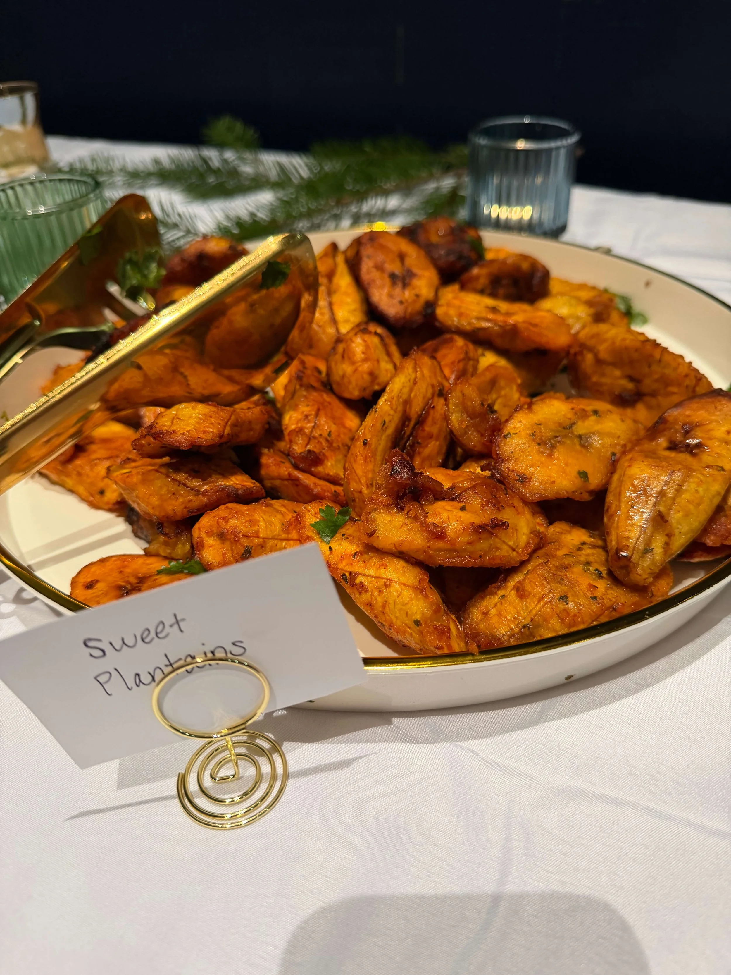 A serving dish of roasted plantains and grilled sausage slices on a table with a white tablecloth. A small sign labeled 'Sweet Plantains' is in front of the dish.