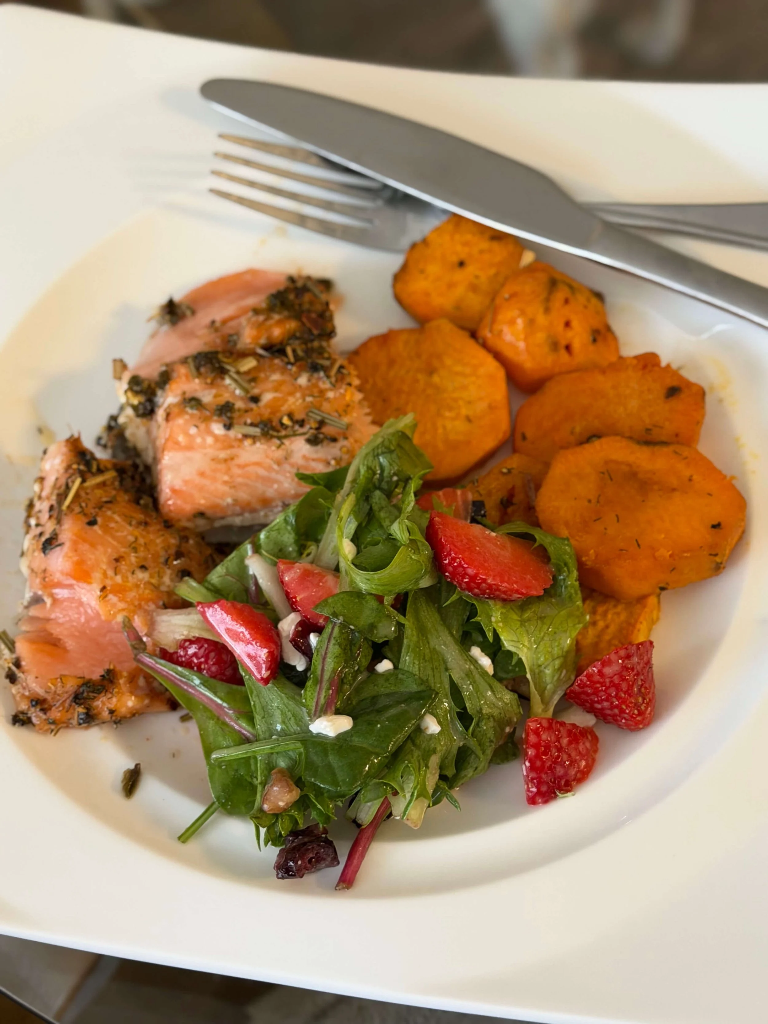 Plate with cooked salmon topped with herbs and black pepper, roasted sweet potatoes, mixed greens salad with strawberries and red onion.