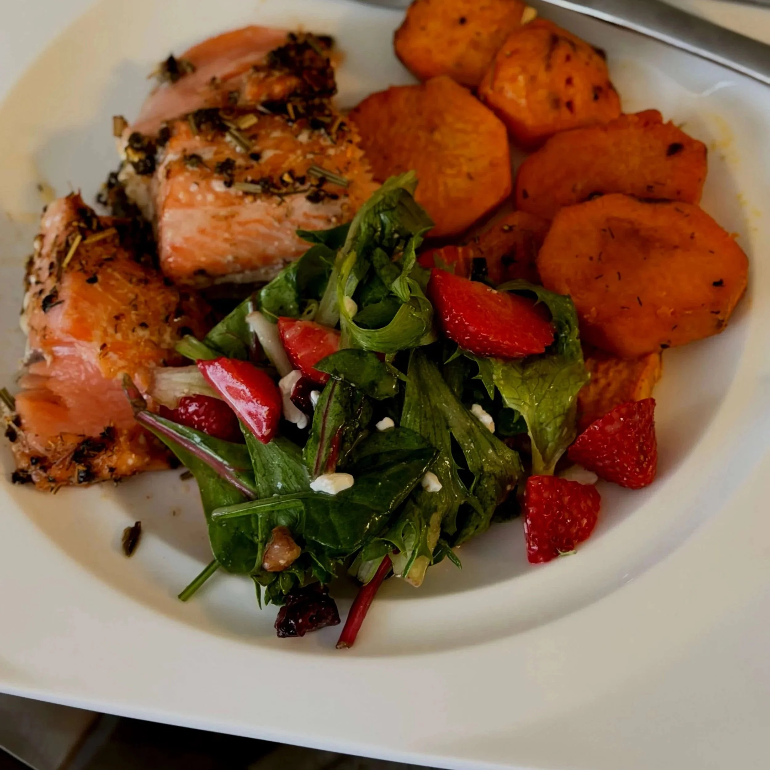 A white plate of roasted salmon, sweet potatoes, a mixed greens salad with strawberries, and herbs.