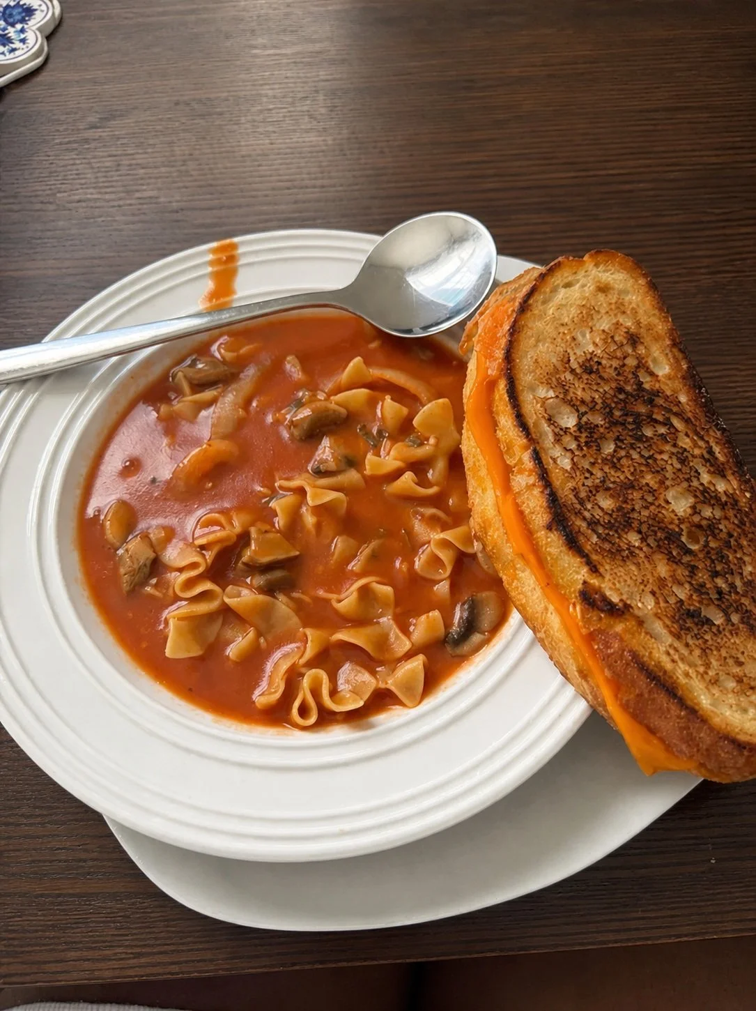 A bowl of tomato-based vegetable soup with pasta, served with a grilled cheese sandwich on the side.