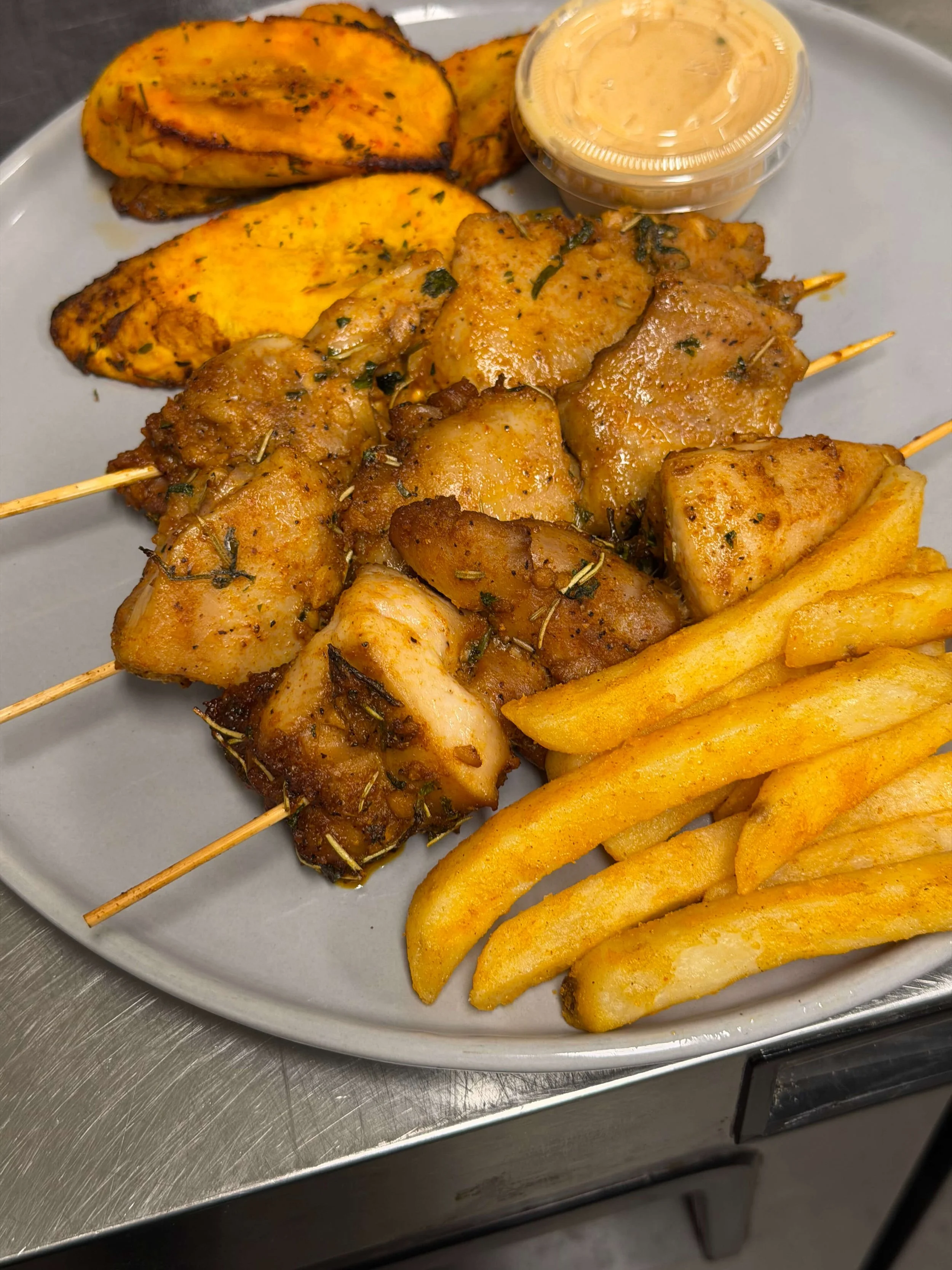 A plate of grilled chicken skewers, roasted plantains, French fries, and a cup of dipping sauce.