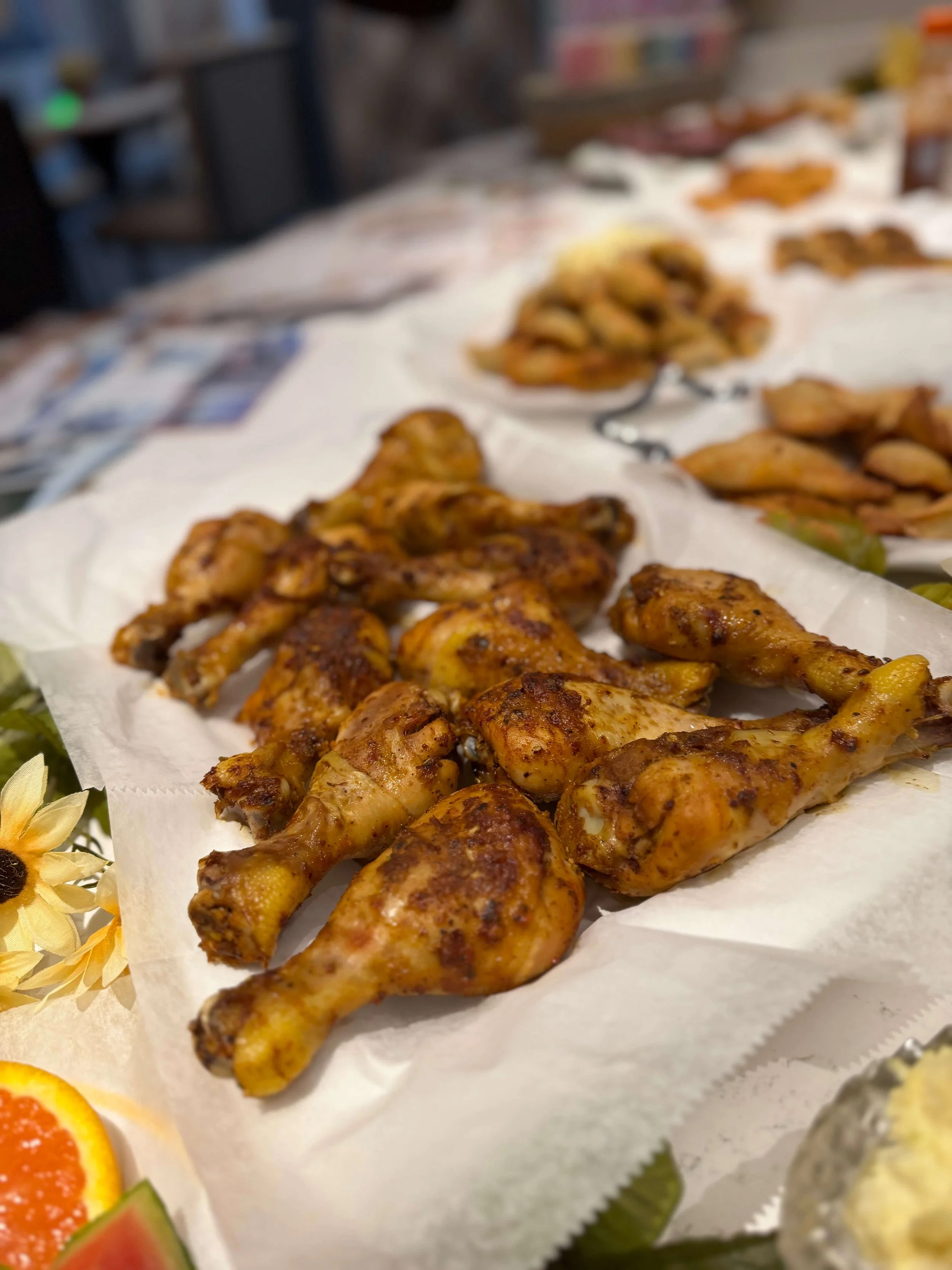 Close-up of grilled chicken drumsticks and wings on a white paper towel, with a blurred background of other food items and a grocery store setting.