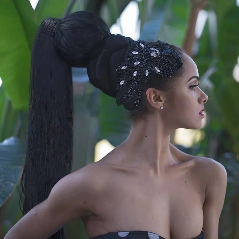 Profile of a woman with an elaborate high ponytail hairstyle, wearing a black headscarf with decorative embellishments, set against a background of green tropical leaves.