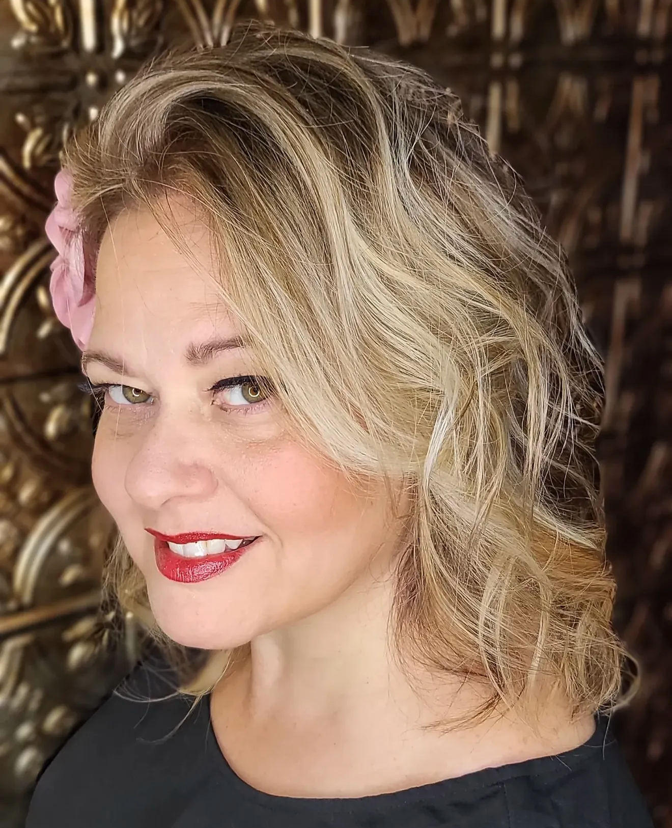A woman with blonde, curly hair styled in loose waves, wearing red lipstick, smiling with light green eyes, and a pink flower hair accessory. The background consists of a decorative wooden wall.