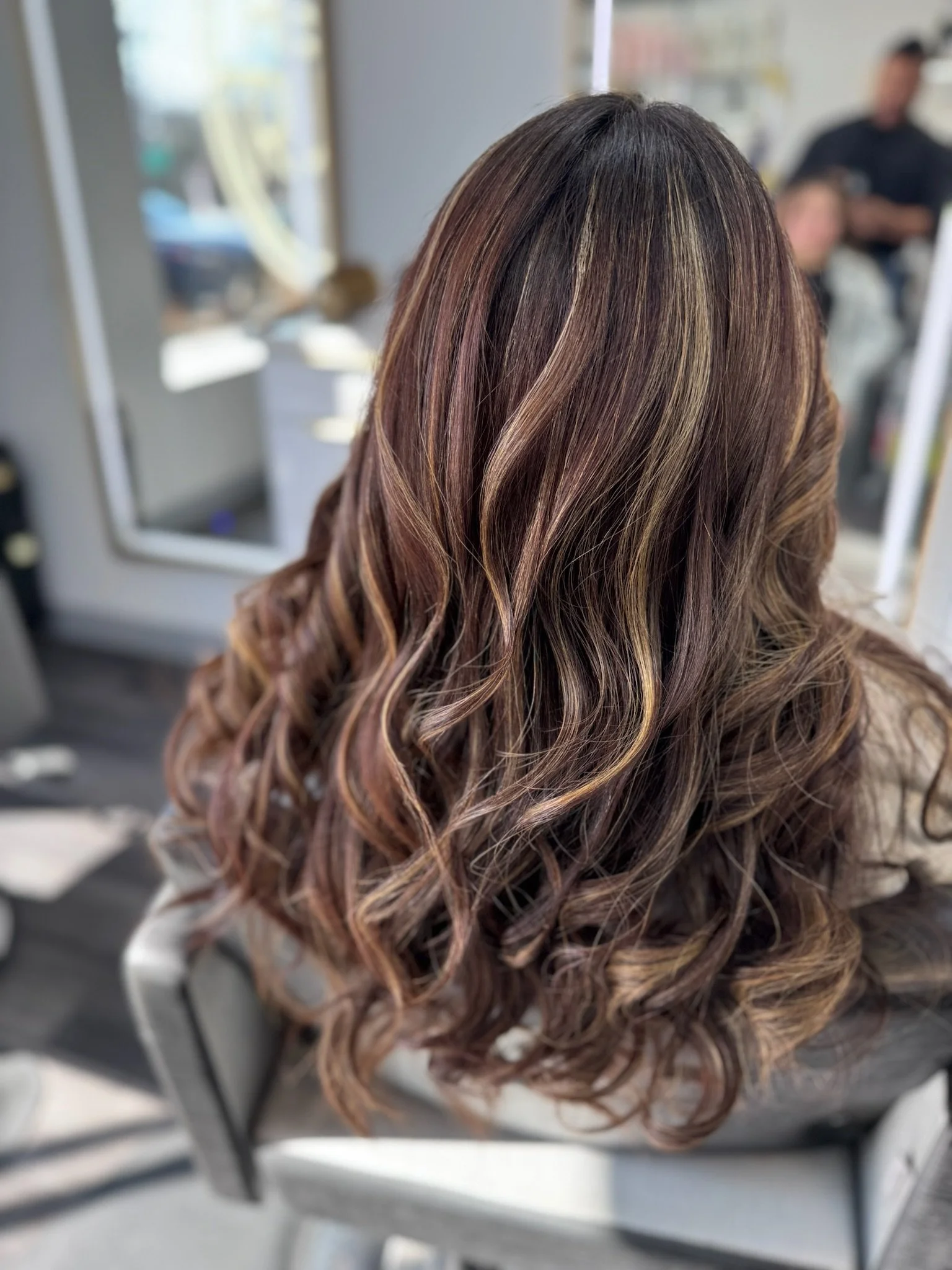 Close-up of a woman's long, wavy hair with highlights in shades of brown, gold, and auburn, styled in loose curls.