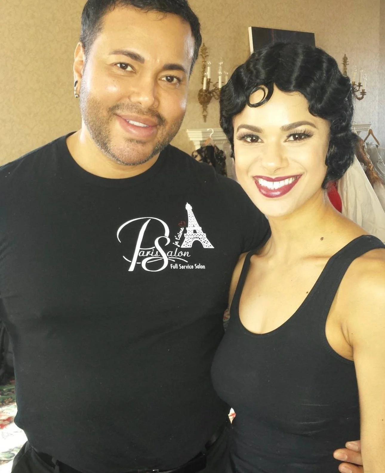 A man and woman smiling and posing for a photo indoors. The man is wearing a black t-shirt with a logo that says 'Paris Salon' and features the Eiffel Tower. The woman has short, styled black hair and is wearing a black sleeveless top.