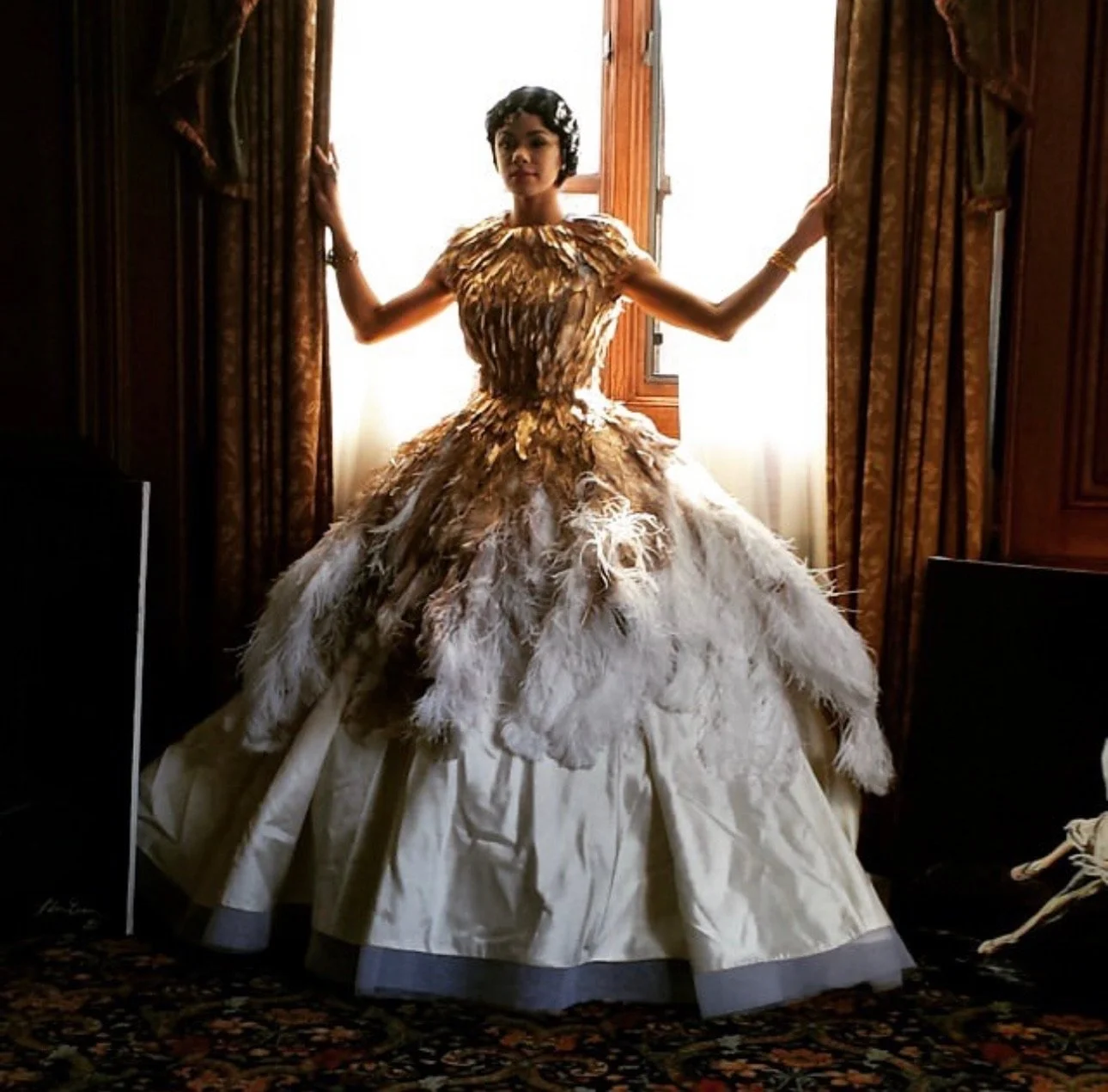 A woman standing indoors in front of a window, wearing an elaborate dress made of feathers and fabric, with her arms extended to the sides, holding open curtains.