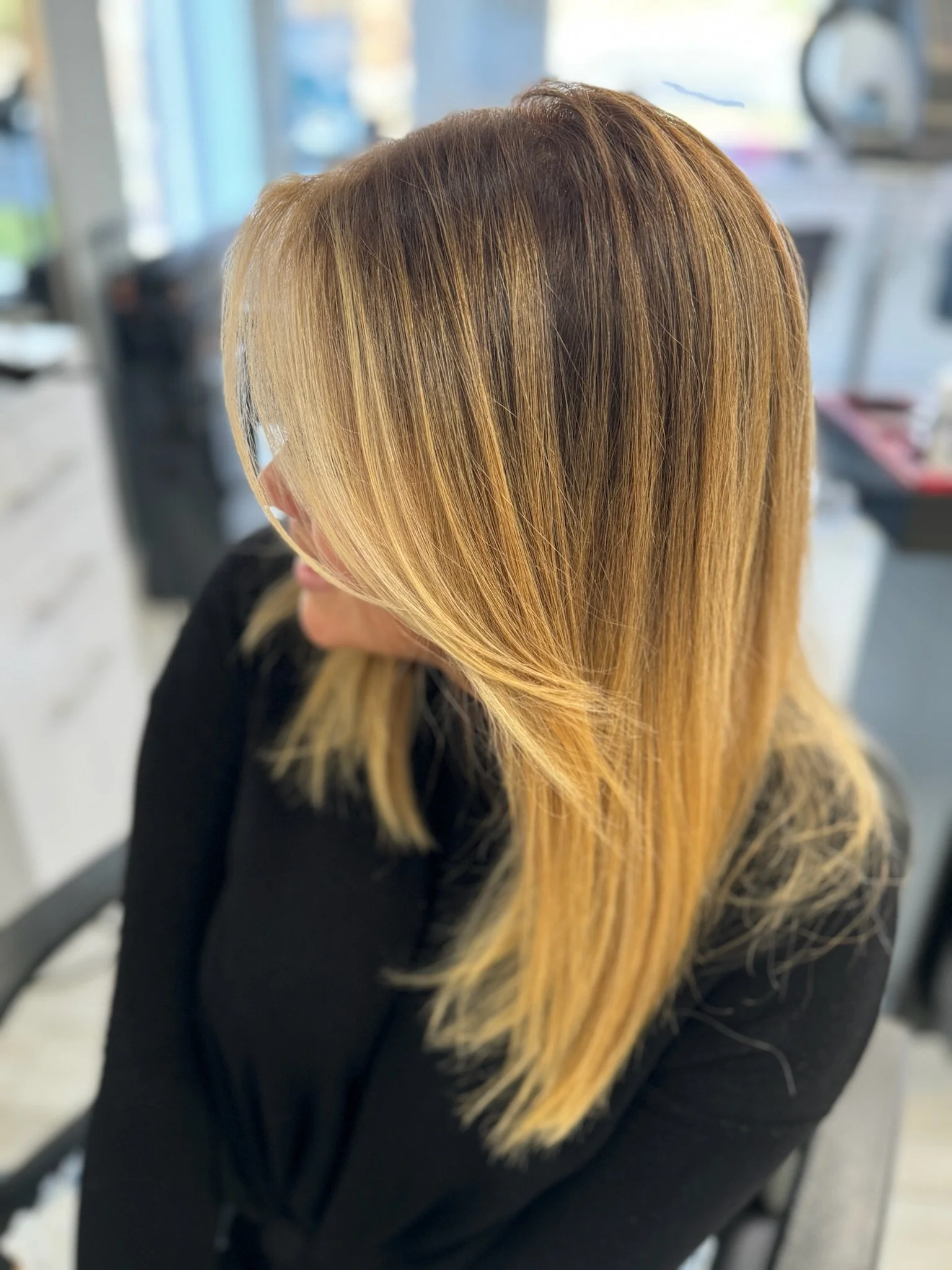 Close-up of a person's hair with blonde highlights, styled straight and parted to the side, in a salon setting.