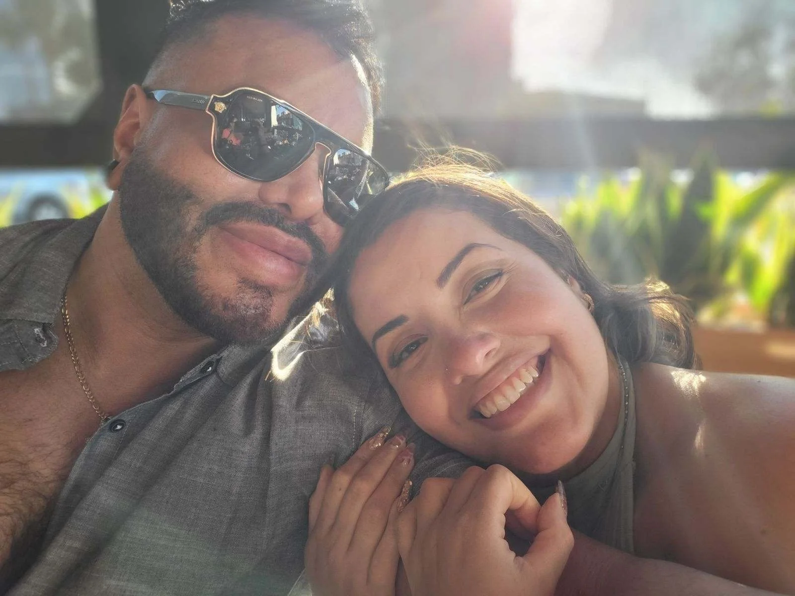 A smiling woman with dark hair and a man with beard and sunglasses, both with faces pressed close together, taking a selfie in a bright, outdoor setting.