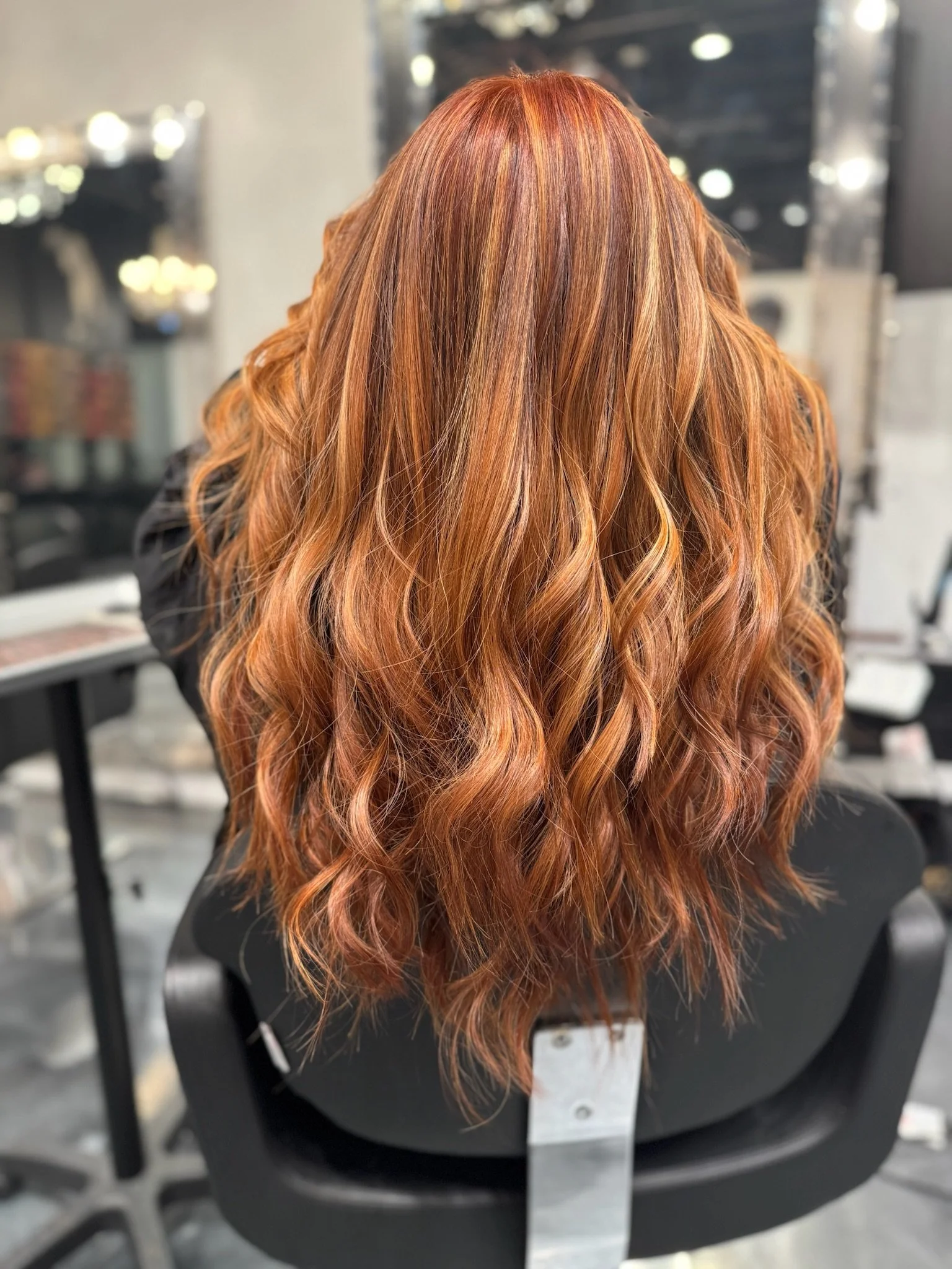 A wig with long, wavy hair in shades of copper, orange, and blonde, displayed on a black mannequin head in a hair salon.