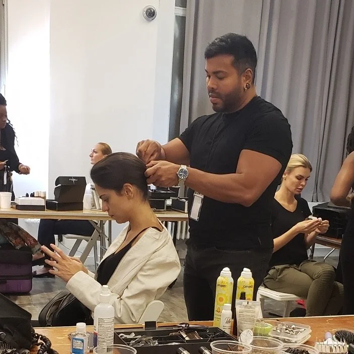 A makeup artist styles a woman's hair at a salon, with other women waiting in the background.