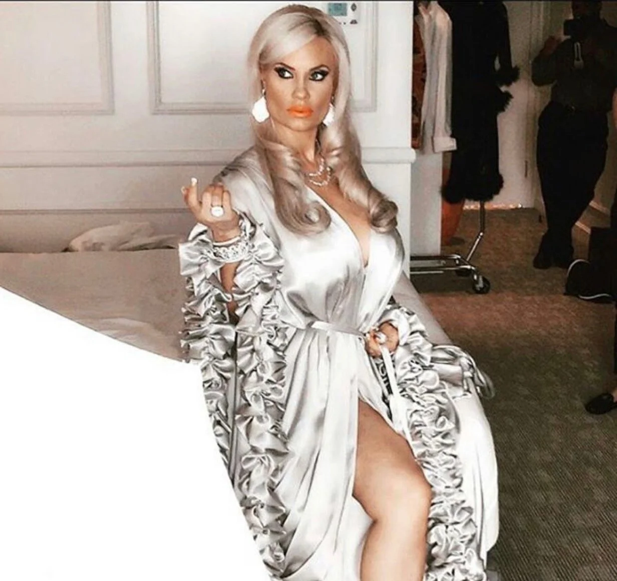 A woman with long, styled blonde hair is sitting on a bed, wearing a shiny, satin pajama set with ruffled details. She has bold makeup, large earrings, and layered jewelry. The woman is holding a cigarette in her right hand and has her left hand resting on her lap, with one leg slightly exposed. The background shows a white paneled wall, a person in black sitting in the distance, and a clothing rack.