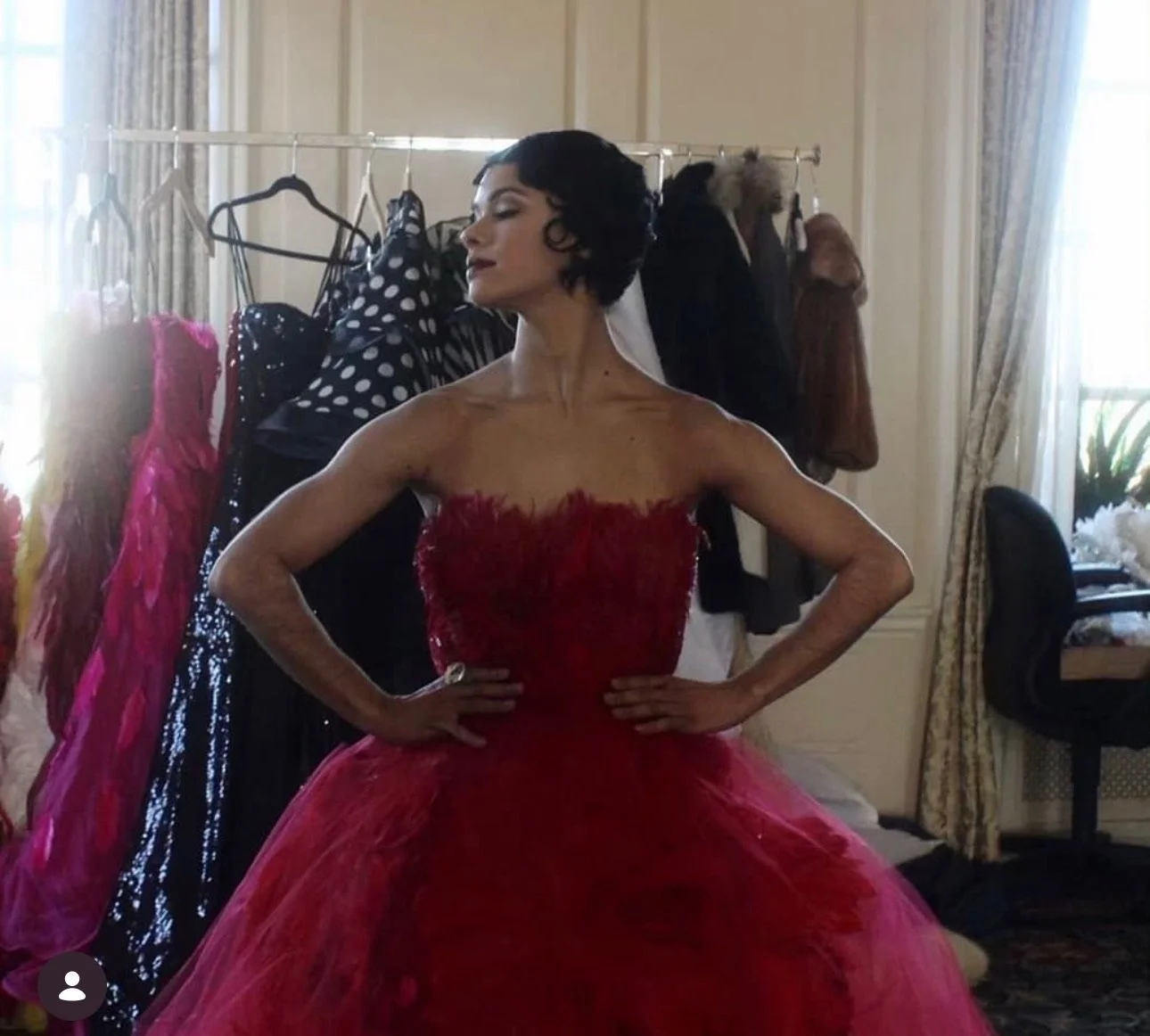 A woman in a red strapless gown with feather details standing in a room with clothing racks and a window with curtains.
