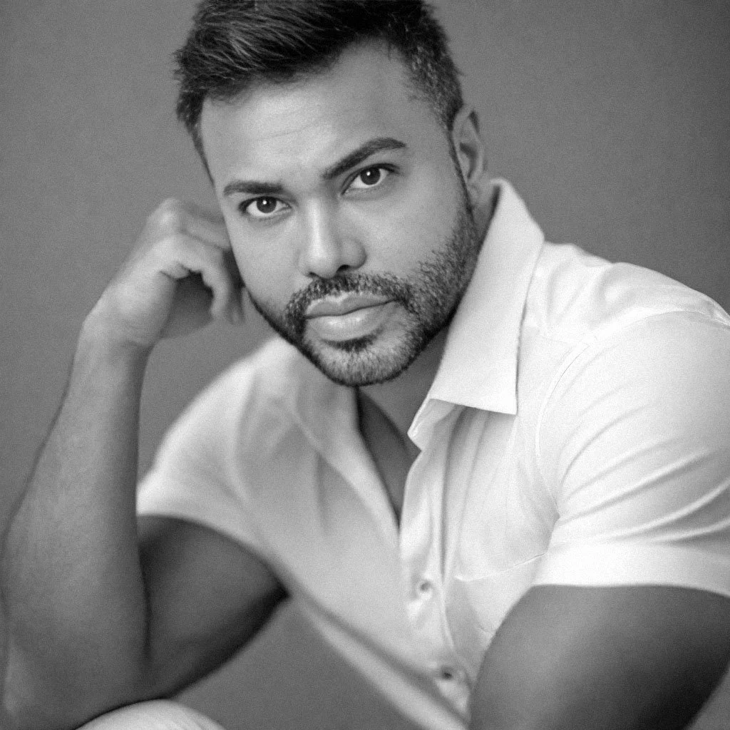 A black-and-white portrait of a man with short hair and a beard, resting his head on his hand, wearing a light-colored shirt, looking directly at the camera with a serious expression.