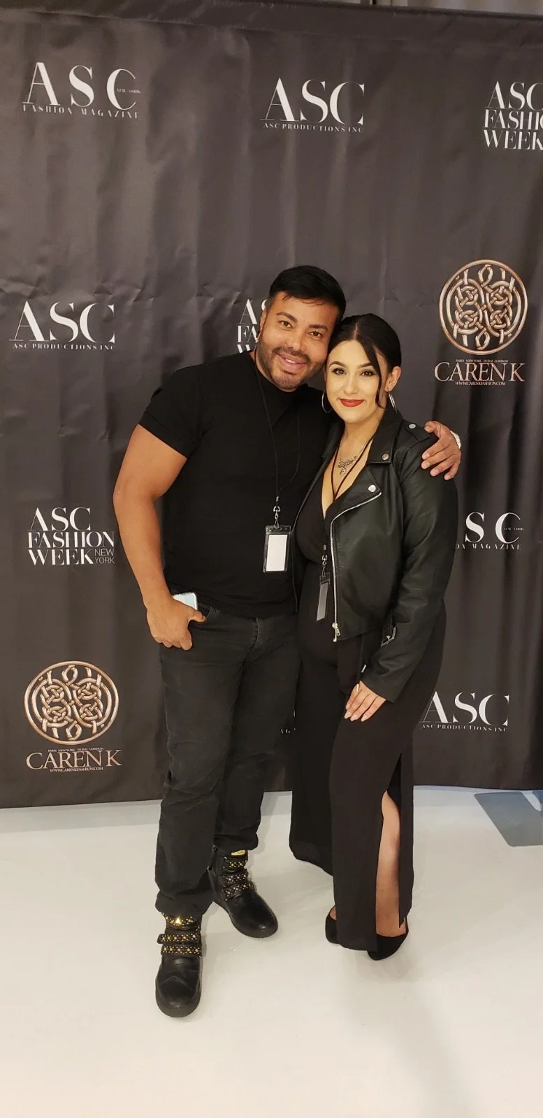 Two people standing together in front of a black backdrop with white logos for ASC Fashion Week and CarenK, smiling and posing for a photo.