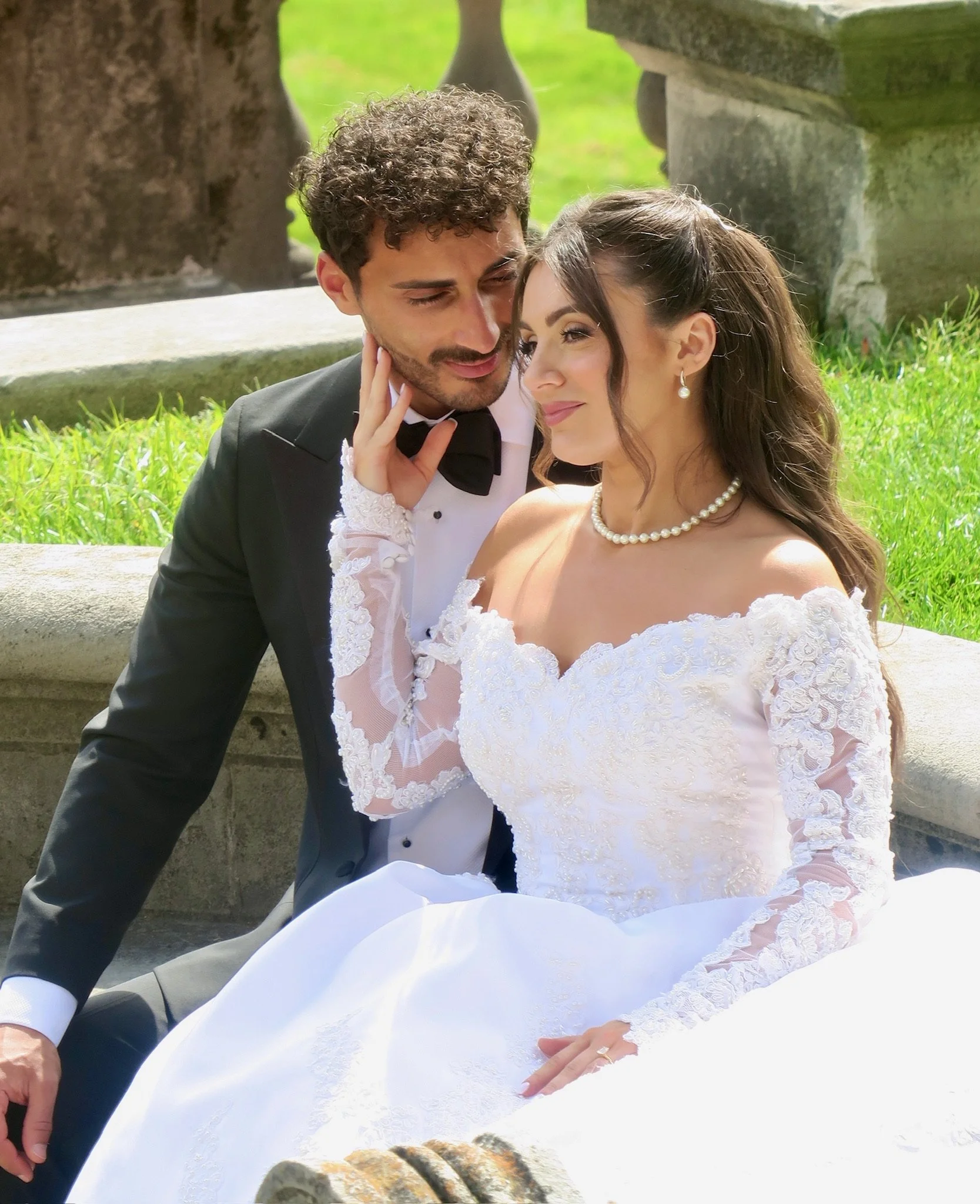 A newlywed couple sitting on stone stairs outdoors, the groom touching the bride’s cheek, surrounded by green grass and stone railing.