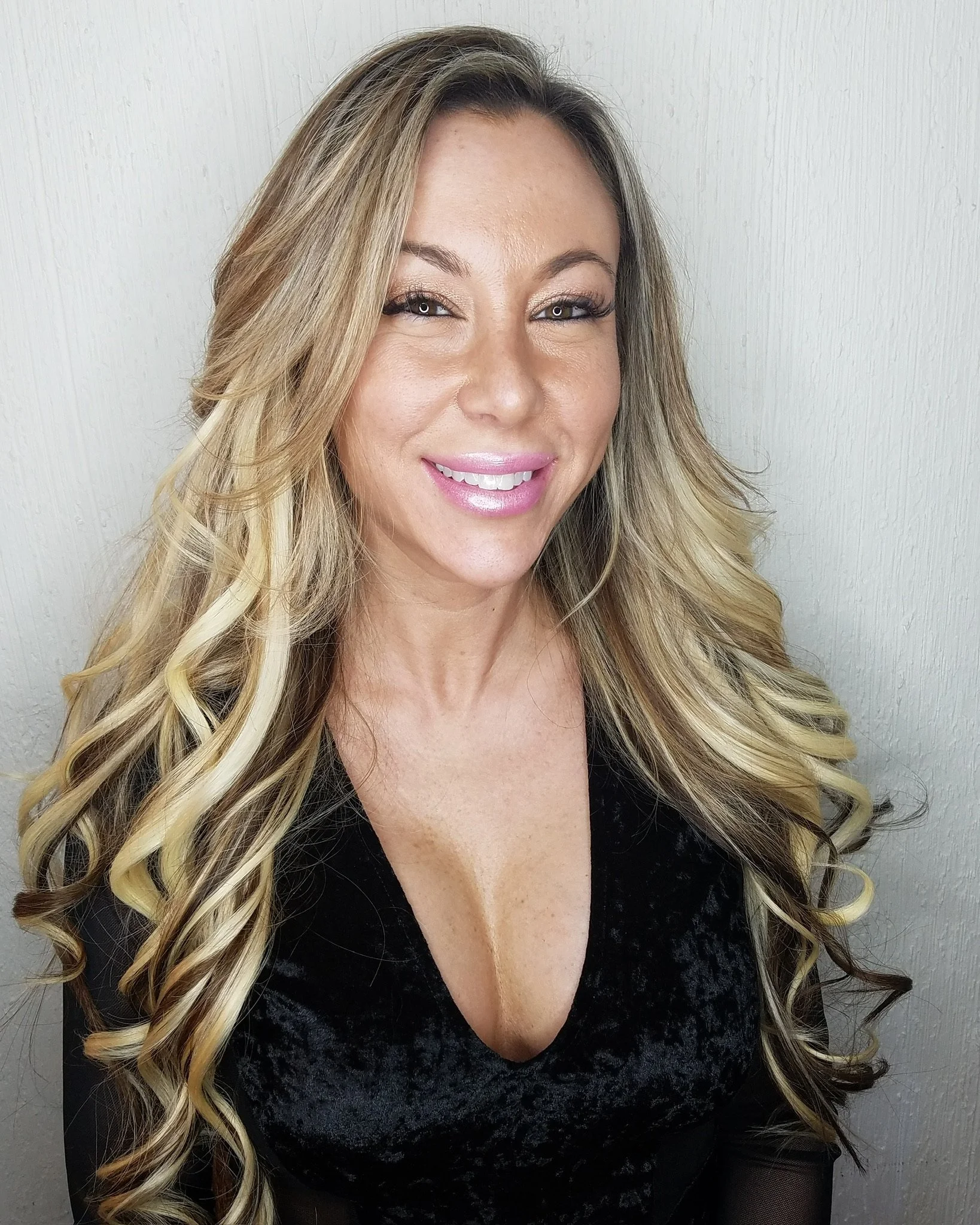 A smiling woman with long, wavy, blonde hair wearing a black velvet dress and makeup against a plain light-colored wall.