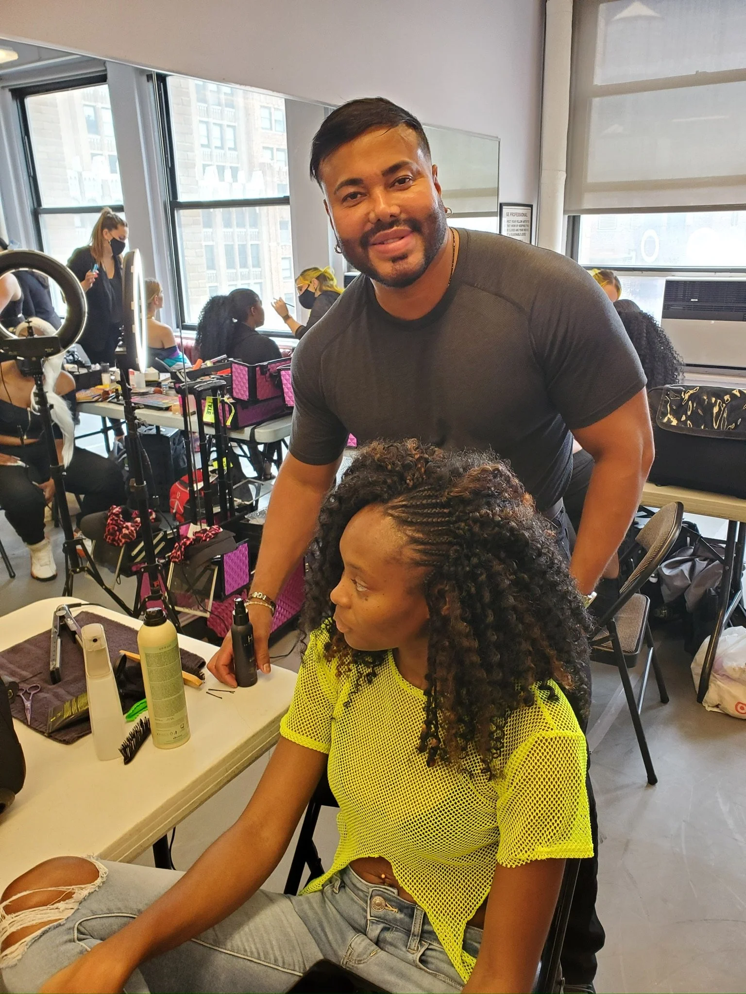 A man standing over a woman with braided hair and a yellow mesh shirt, while other people work at makeup stations in a well-lit room.