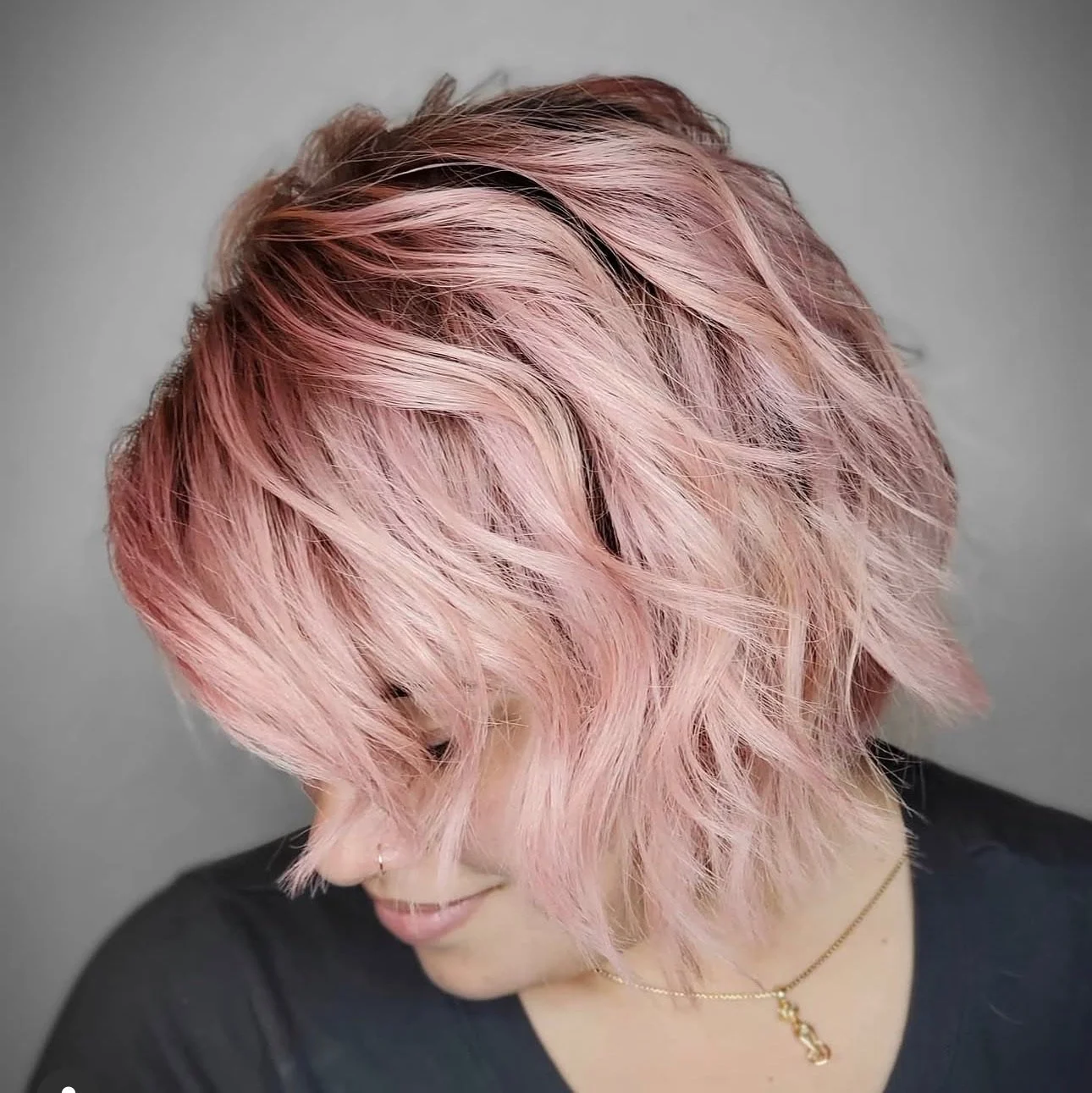 Close-up of a woman with pink, wavy, layered bob haircut, wearing a black top and necklace with a pendant.