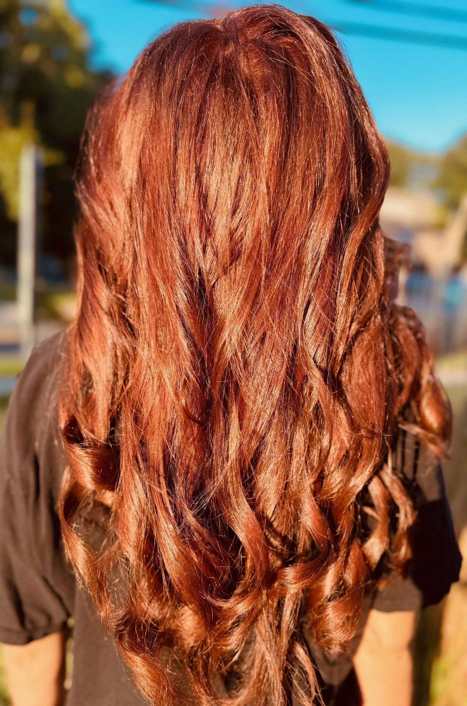 Back of a person with long, wavy, reddish-brown hair wearing a black shirt outdoors on a sunny day.