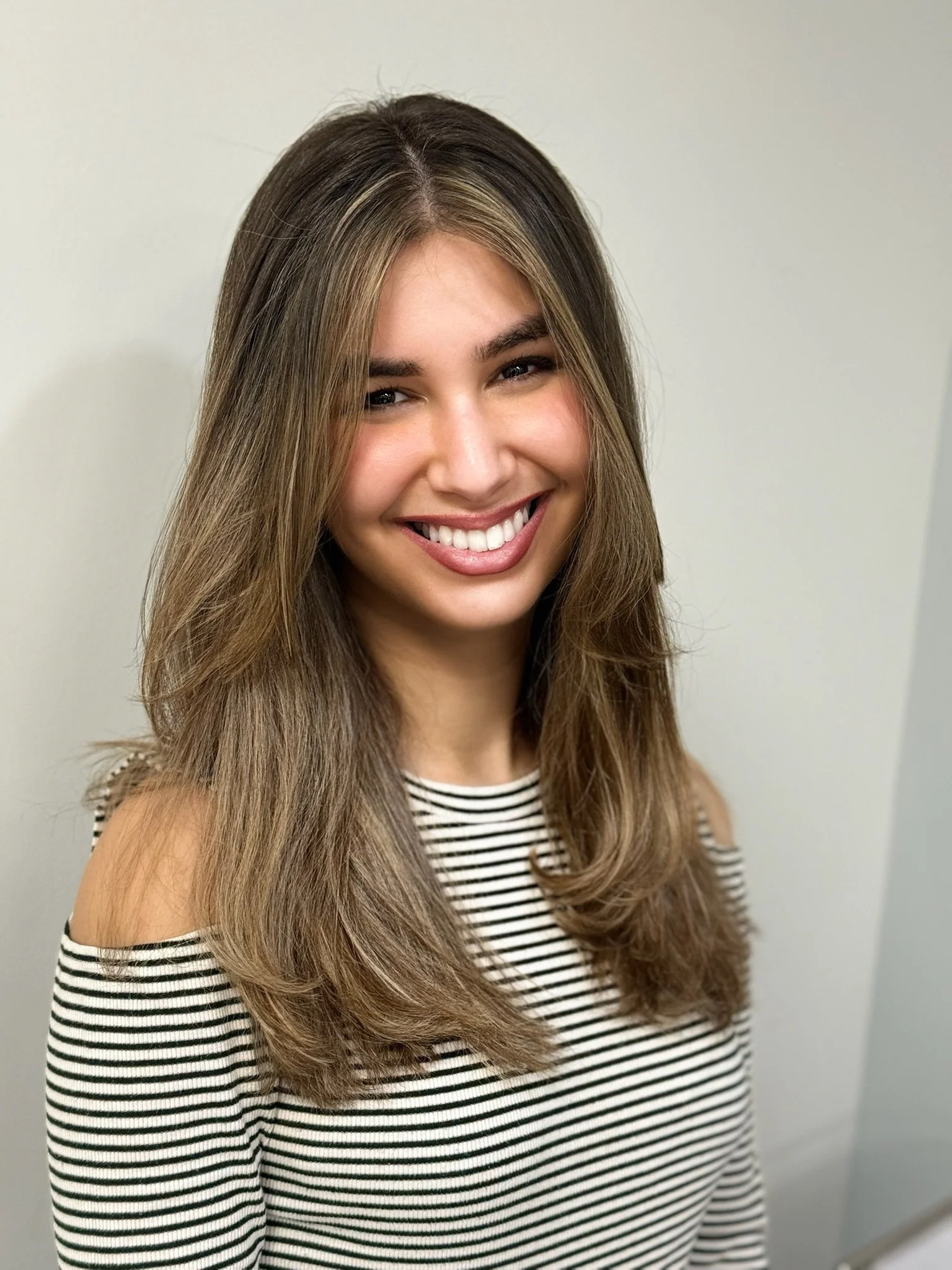 A young woman with long brown hair, smiling, wearing a striped off-shoulder top, standing against a plain light-colored background.
