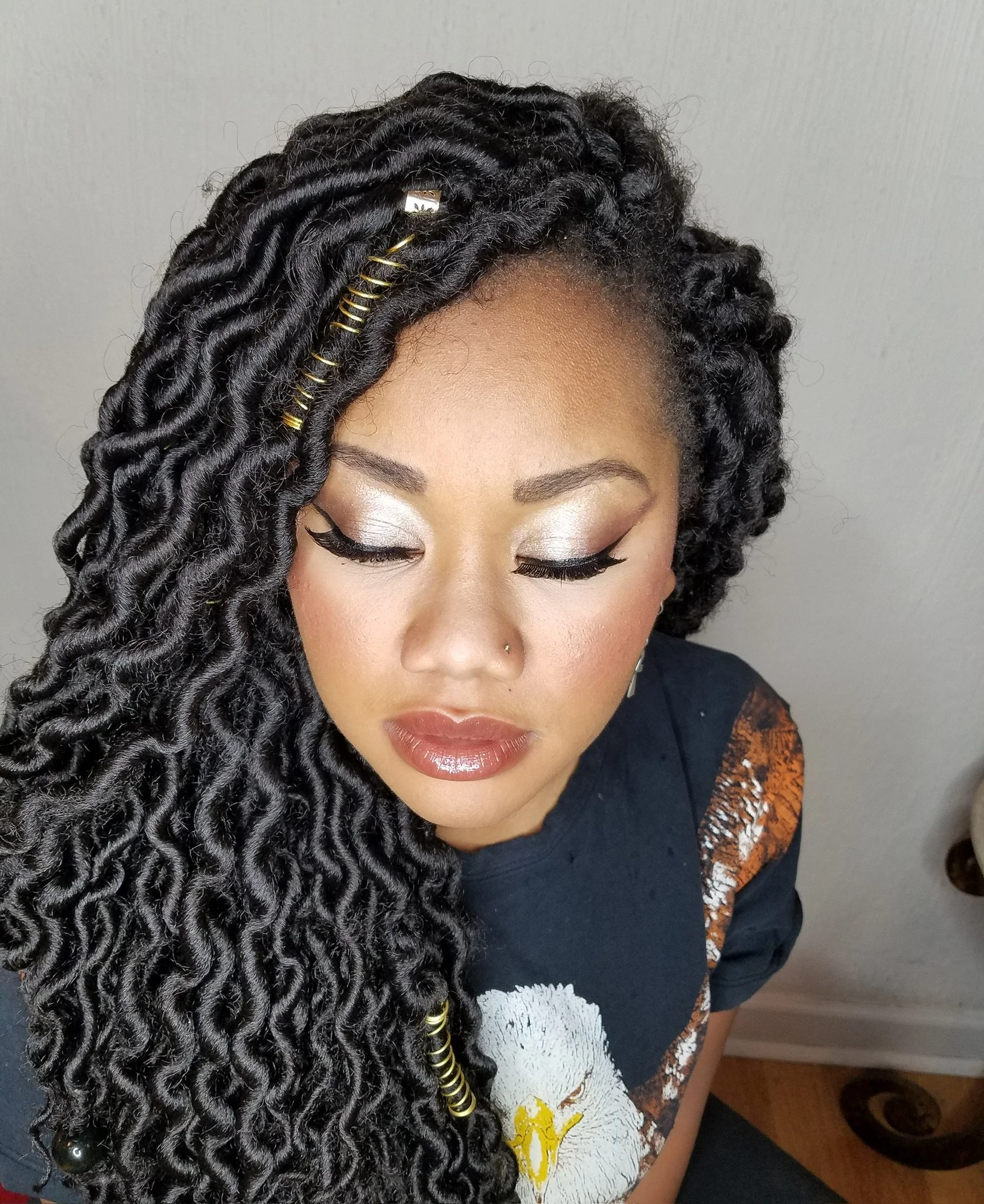 Close-up of a woman with dark, wavy, faux locs styled to one side, decorated with gold hair cuffs and beads, wearing makeup with voluminous eyelashes and glossy lips, dressed in a black t-shirt with a white graphic, sitting on a wooden floor.