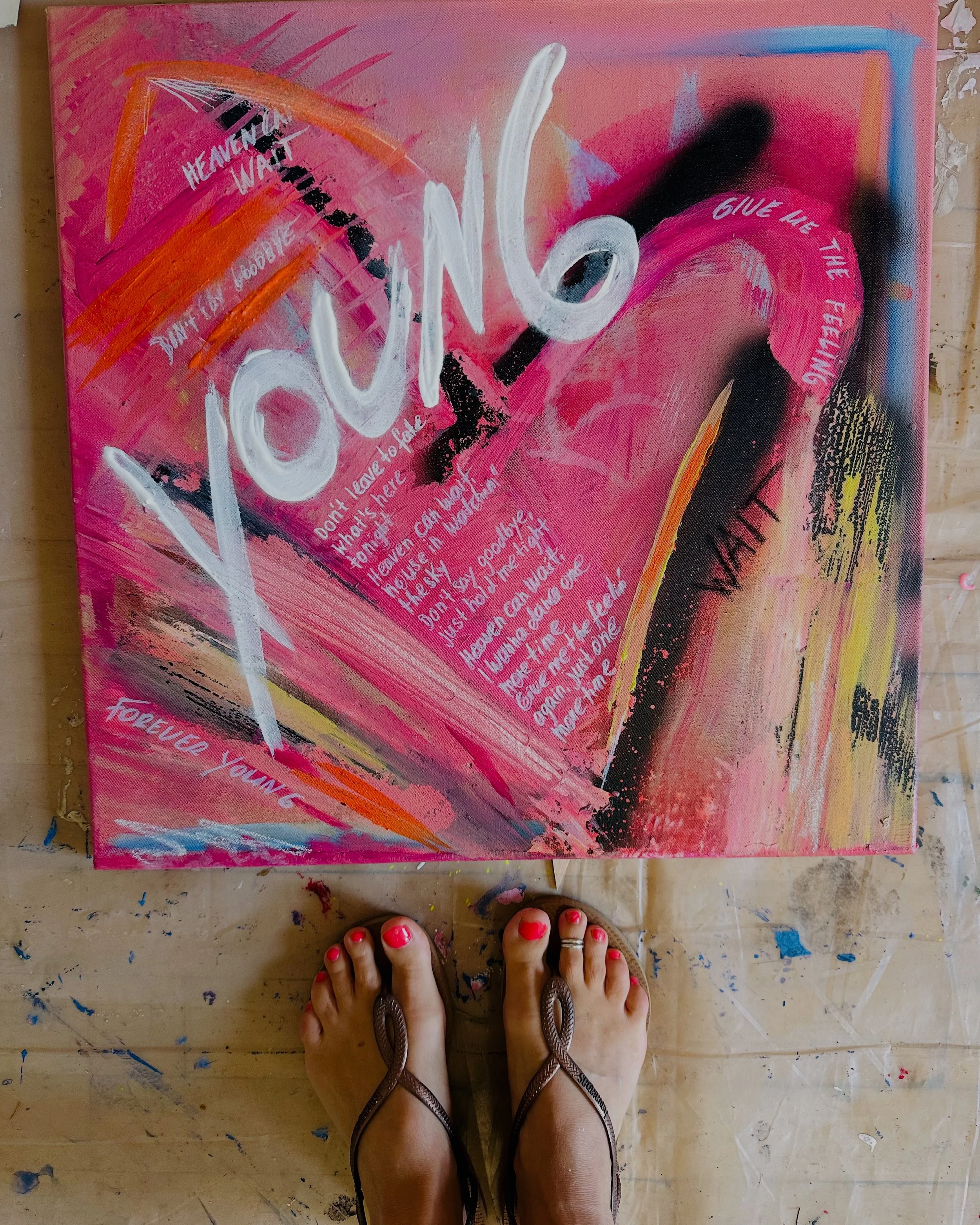 Ein buntes abstraktes Gemälde mit Worten, die wie Graffiti aussehen, in Rosa, Schwarz, Weiß, Orange und Blau. Ein Paar Füße mit roten Lackierten Nägeln und Sandalen steht auf einem bunten, undekorierten Boden.