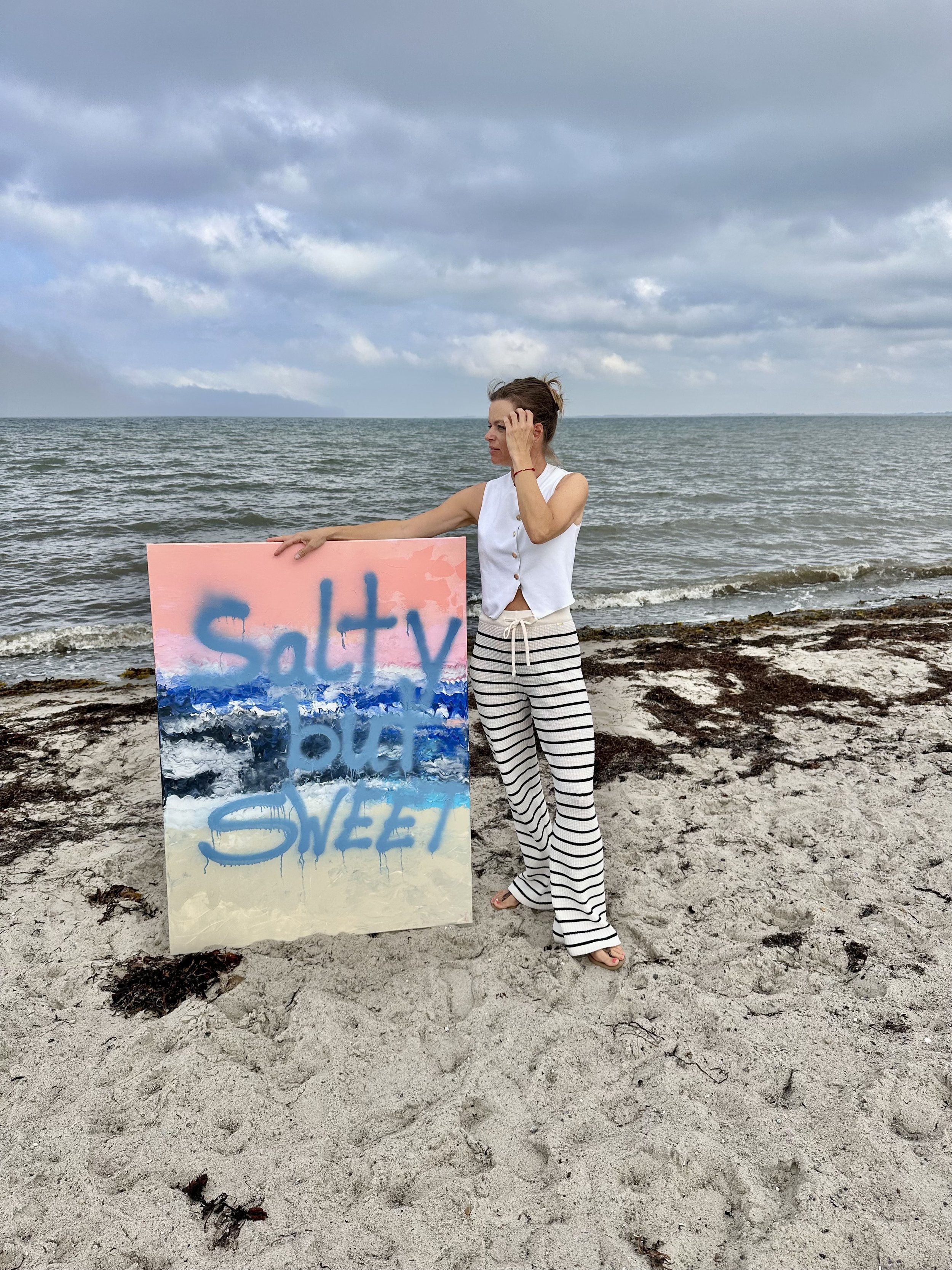 Frau am Strand hält ein bunt bemaltes Schild mit der Aufschrift " Sultry & Sweg". Sie trägt ein weißes Top und schwarze-weiß gestreifte Hosen. Der Himmel ist bewölkt, das Wasser ist ruhig, und der Strand ist mit Seegras bedeckt.