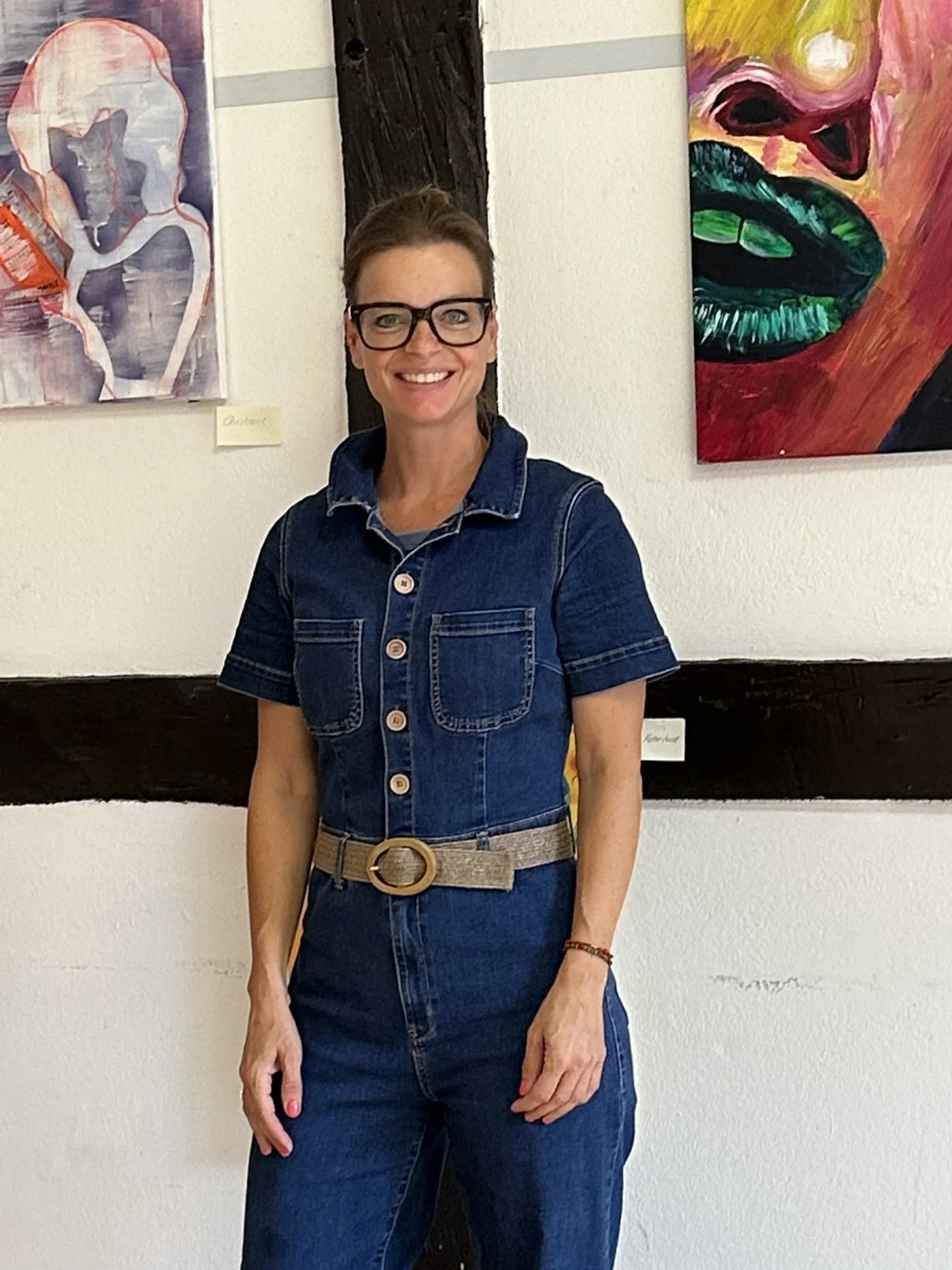 Eine Frau mit Brille in Jeanskleidung steht vor einer weißen Wand mit mehreren bunten Gemälden.