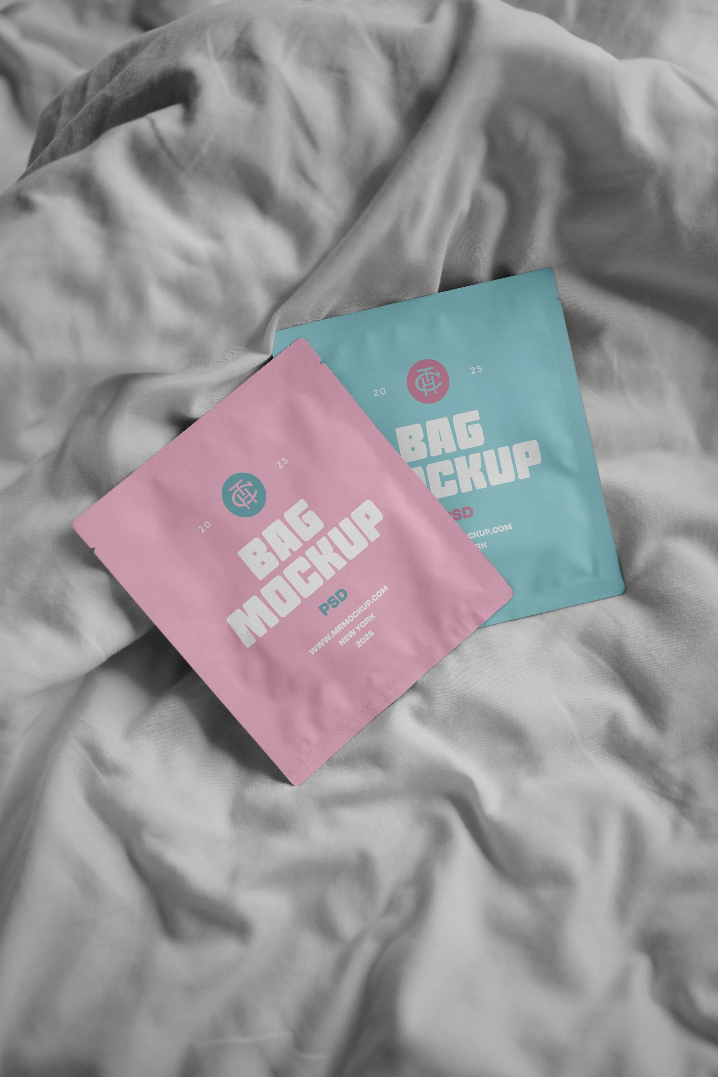 Two small, pastel-colored pocket-sized packages labeled "BAG MOCKUP PSD" in white text, resting on a crumpled, gray bedsheet.