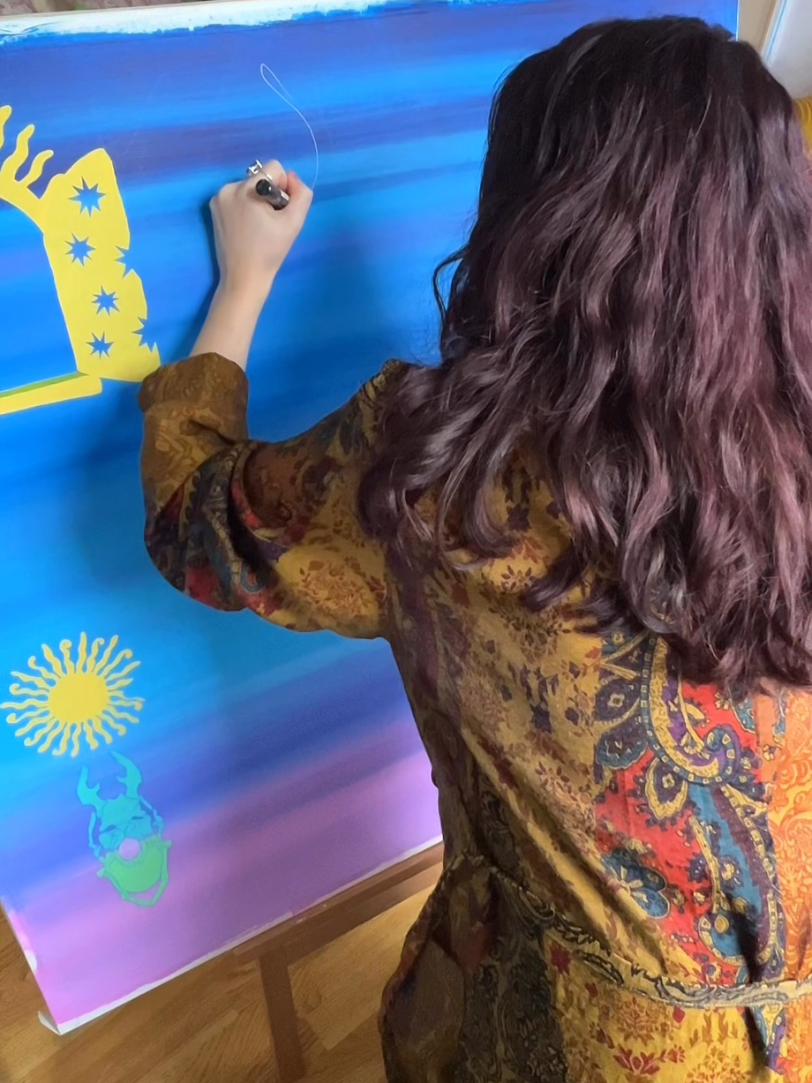 A woman with wavy, reddish-brown hair is drawing on a colorful, gradient background on a large vertical board. She is wearing a patterned, floral-colored long-sleeve top.