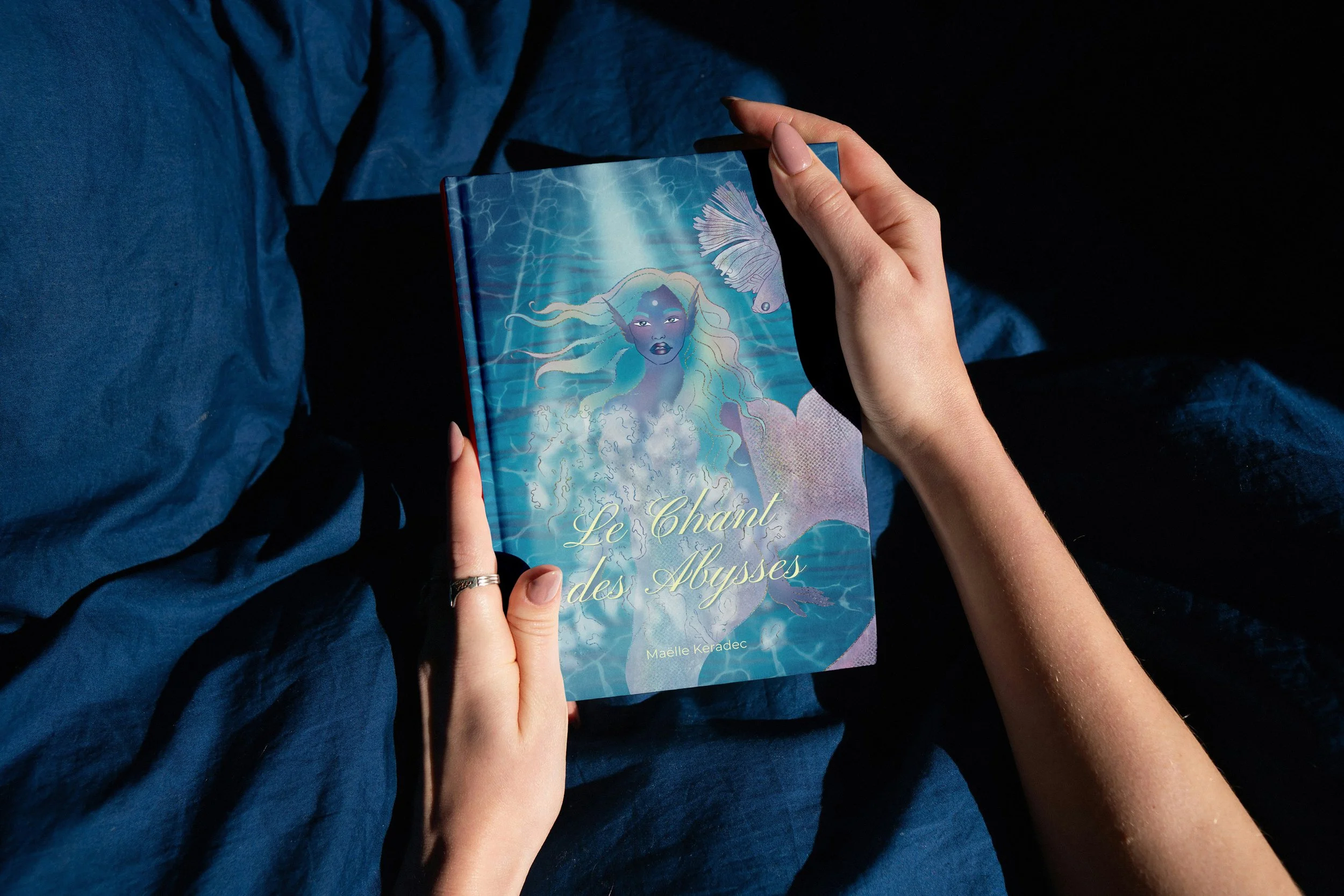 A person with light skin holding a book titled 'Le Chant des Abysses' by Maëlle Keradec, sitting on a blue fabric surface with a dark background.