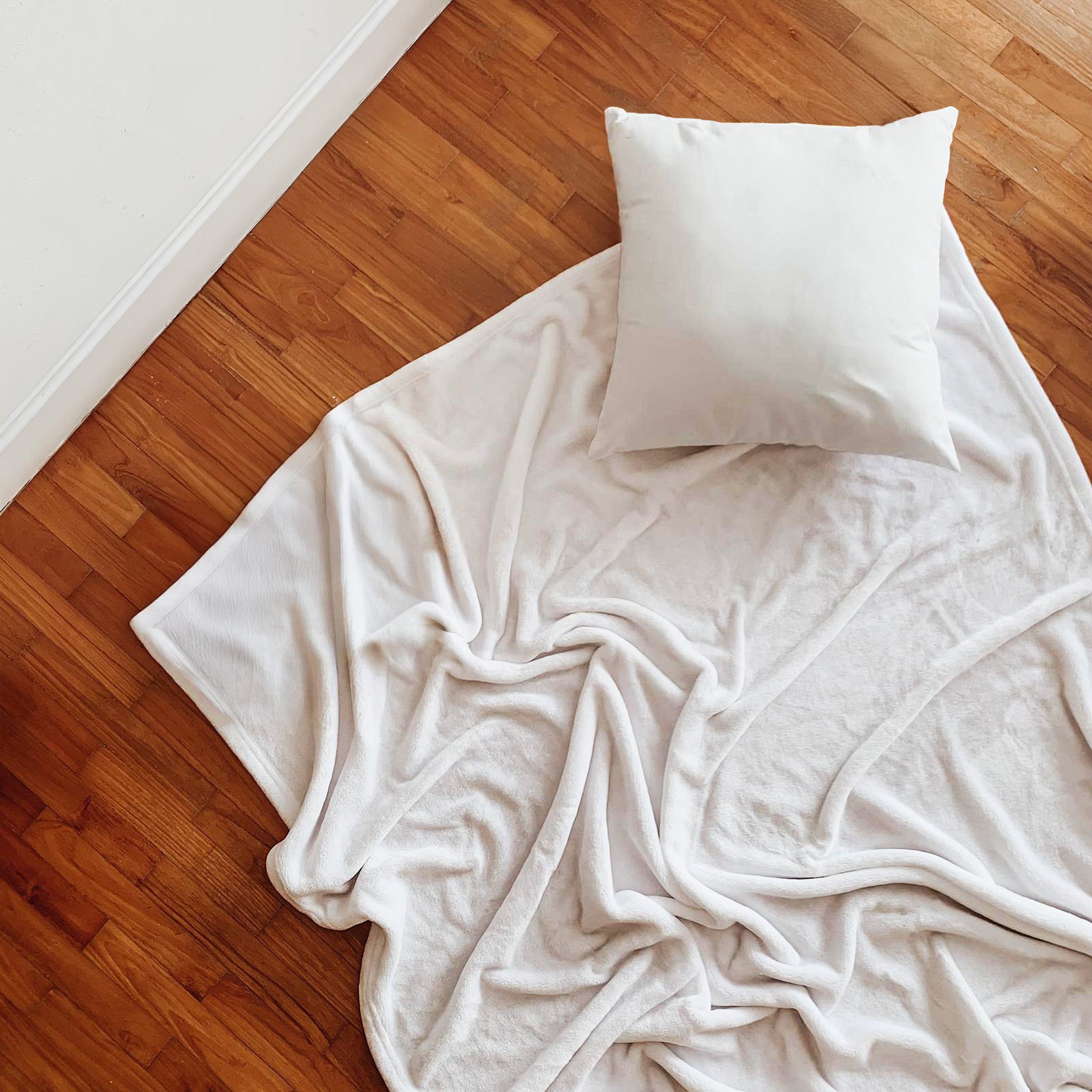 A white pillow resting on a white sheet on a wooden floor near a beige wall with white baseboard.
