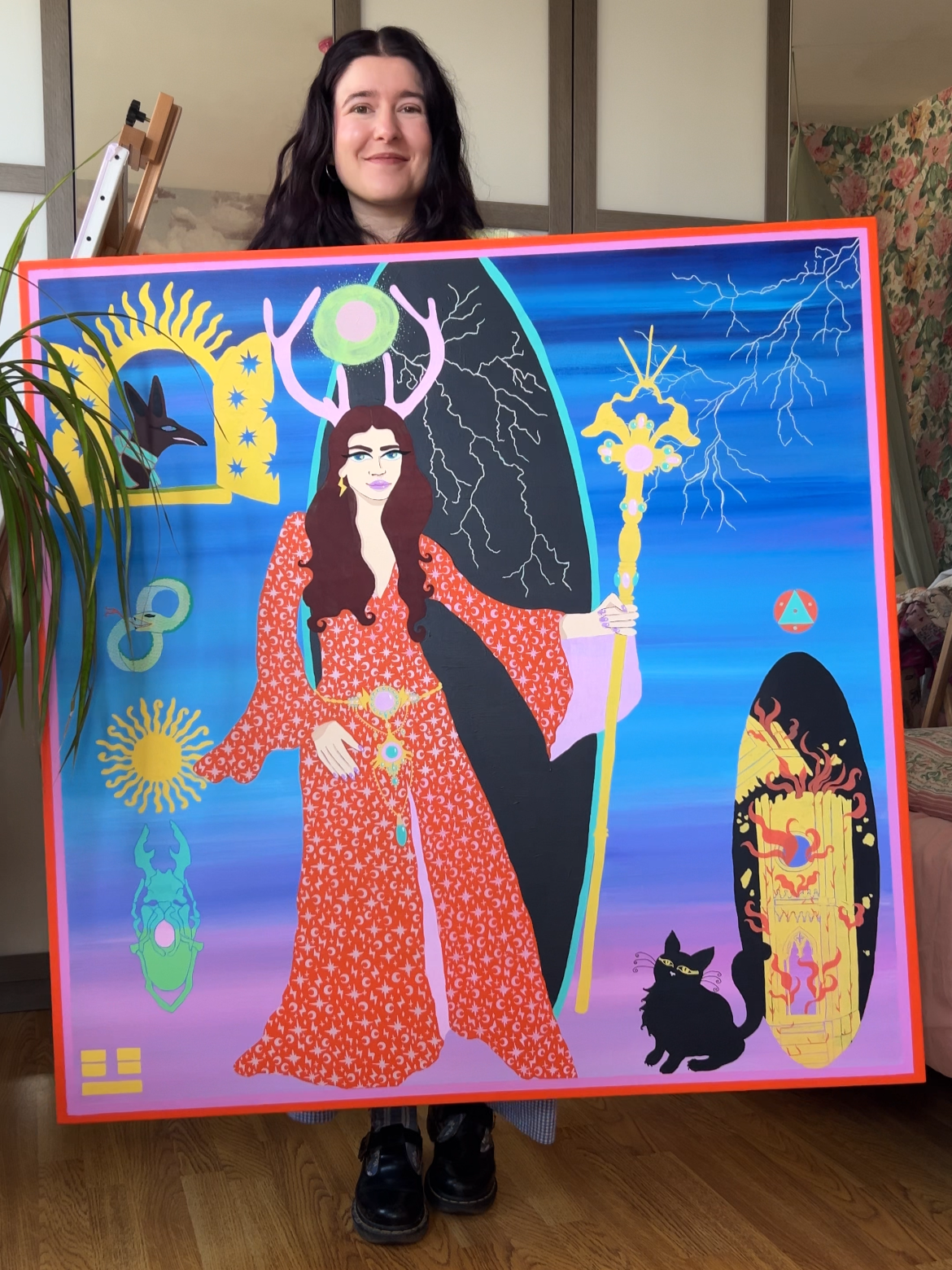 A woman with dark hair holding a colorful, surreal painting that features a mystical female figure with antlers, surrounded by various celestial and symbolic elements, including lightning, a black cat, and abstract designs, set against a vibrant blue background.