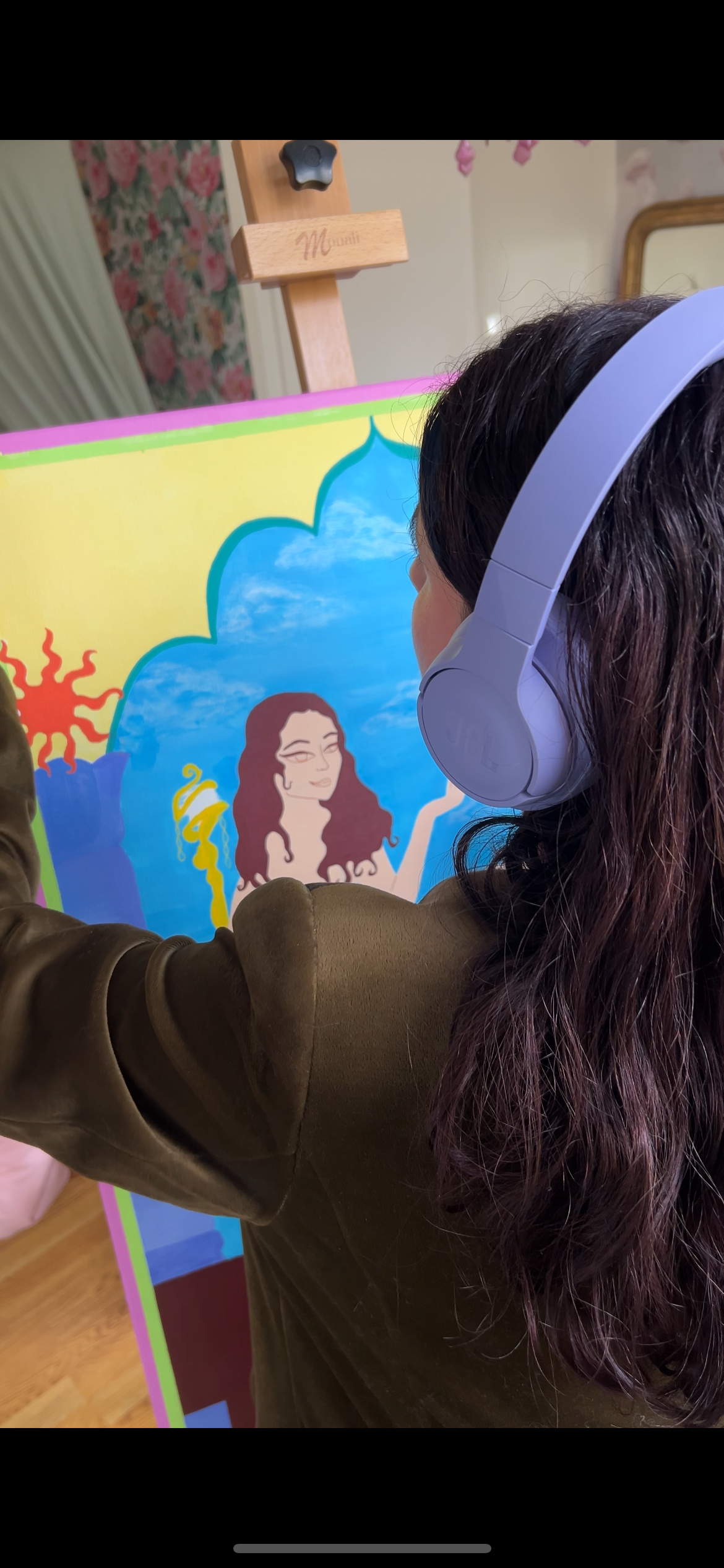 A woman with long dark hair wearing white headphones and a brown jacket looking at a colorful children's illustration on a board.