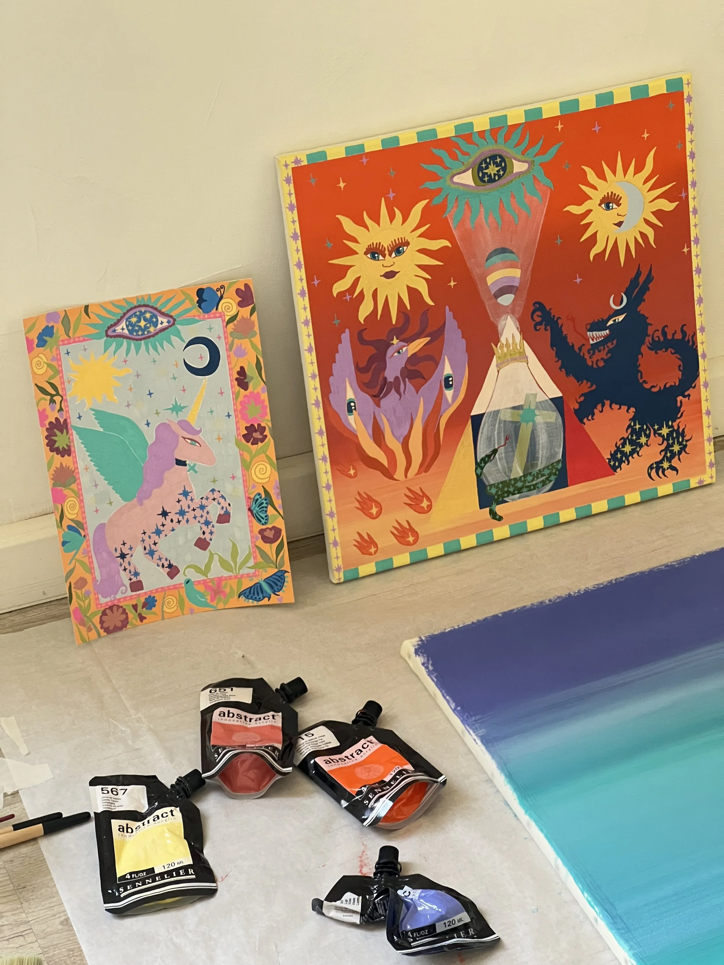 Colorful paintings leaning against a wall and watercolor paint tubes on a surface.
