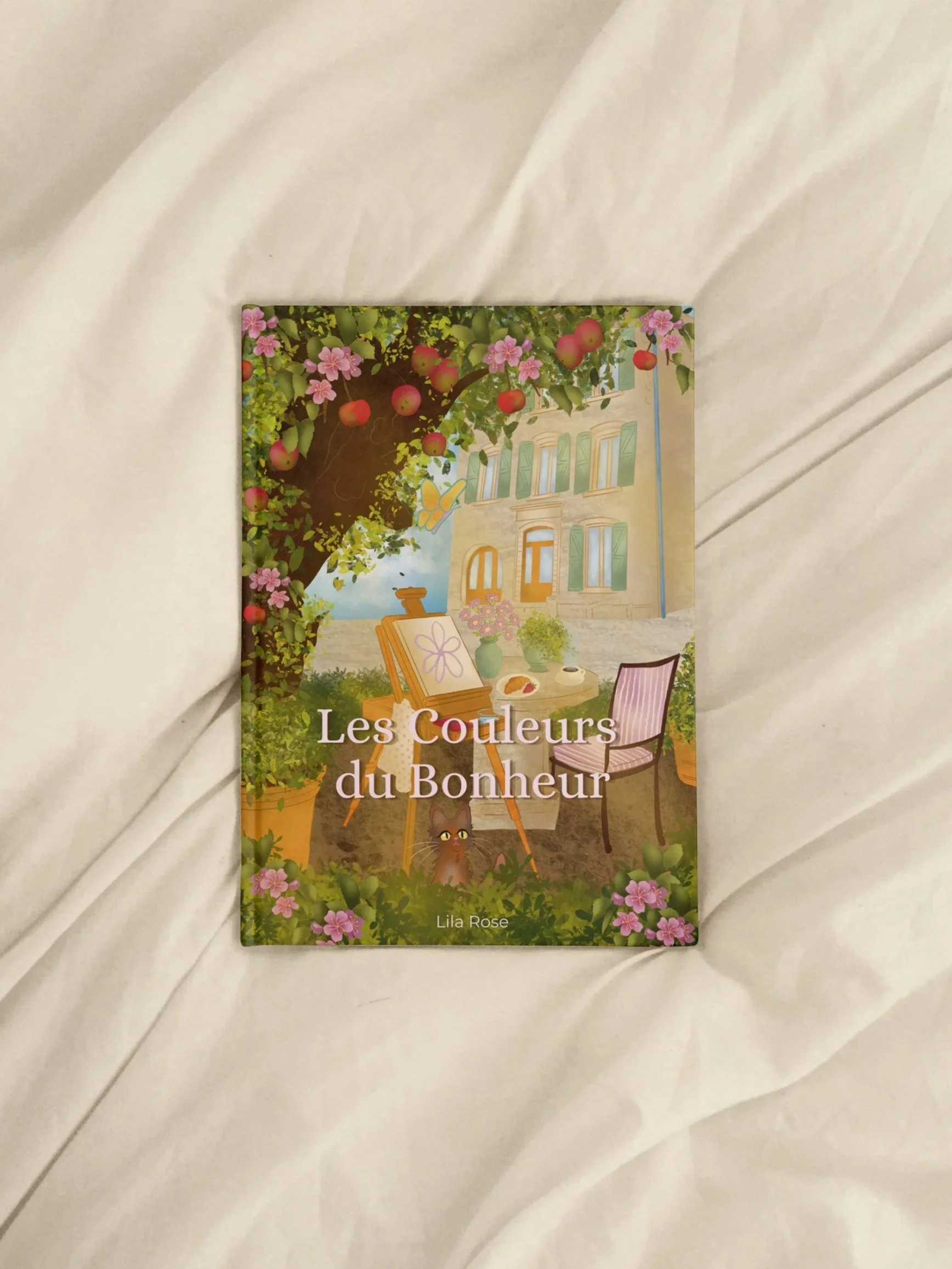 Book titled 'Les Couleurs du Bonheur' by Lila Rose showing a colorful house, apple tree, flowers, and outdoor furniture with a kitten.