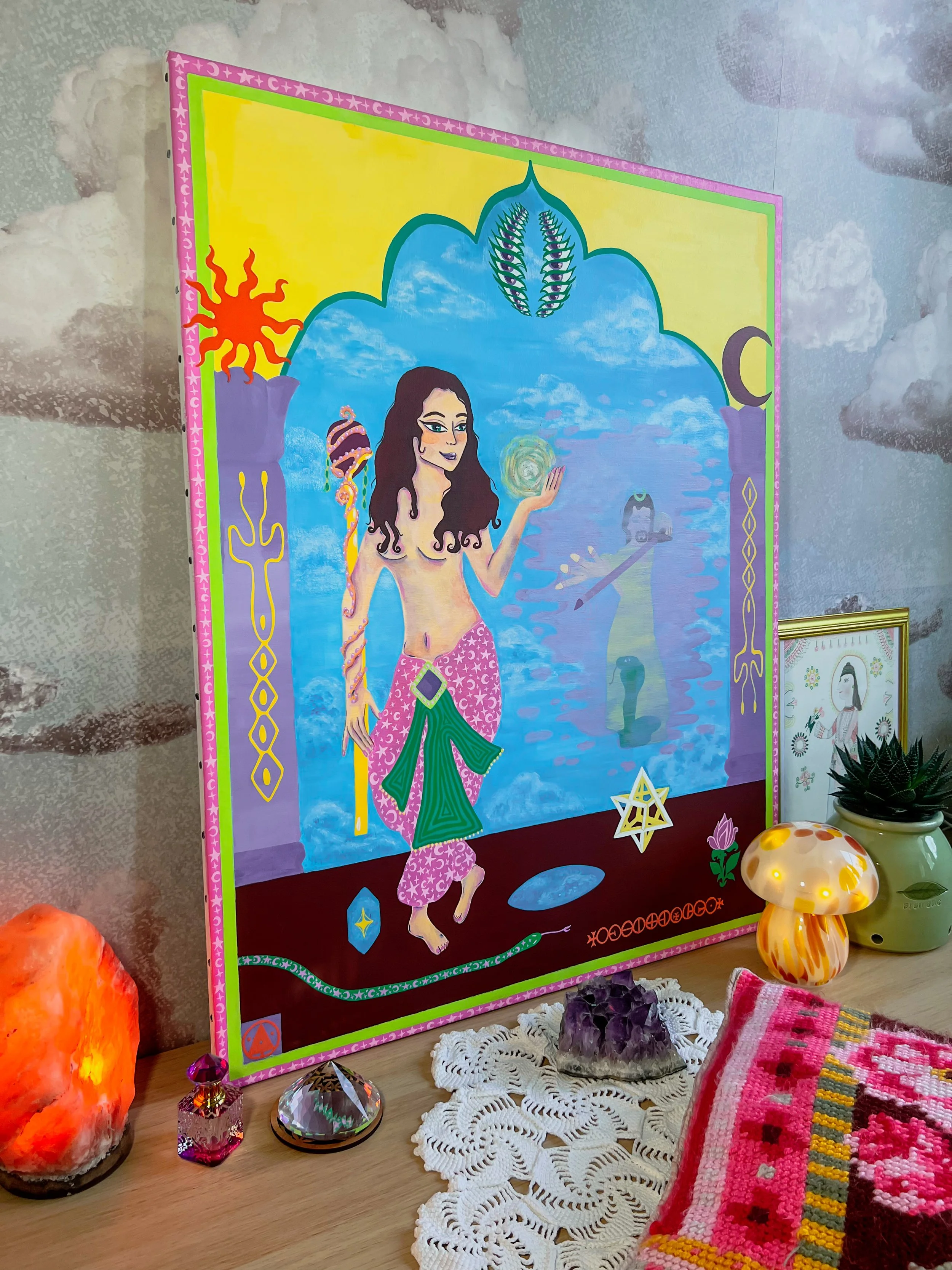 A colorful painting of a woman with long dark hair, fantasy-style background with clouds and celestial symbols, decorative border, placed on a Christmas-like setup with crystals, a salt lamp, and hanging ornaments on a table.