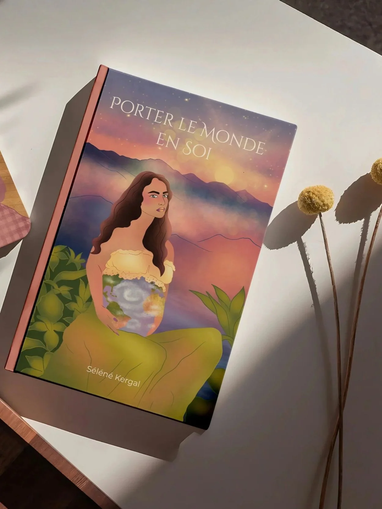 Book titled 'Porter le Monde en soi' by Selené Kergal, with illustrated woman sitting outdoors in a natural landscape at sunset, holding a glowing globe. The background features mountains and a pastel-colored sky with stars.