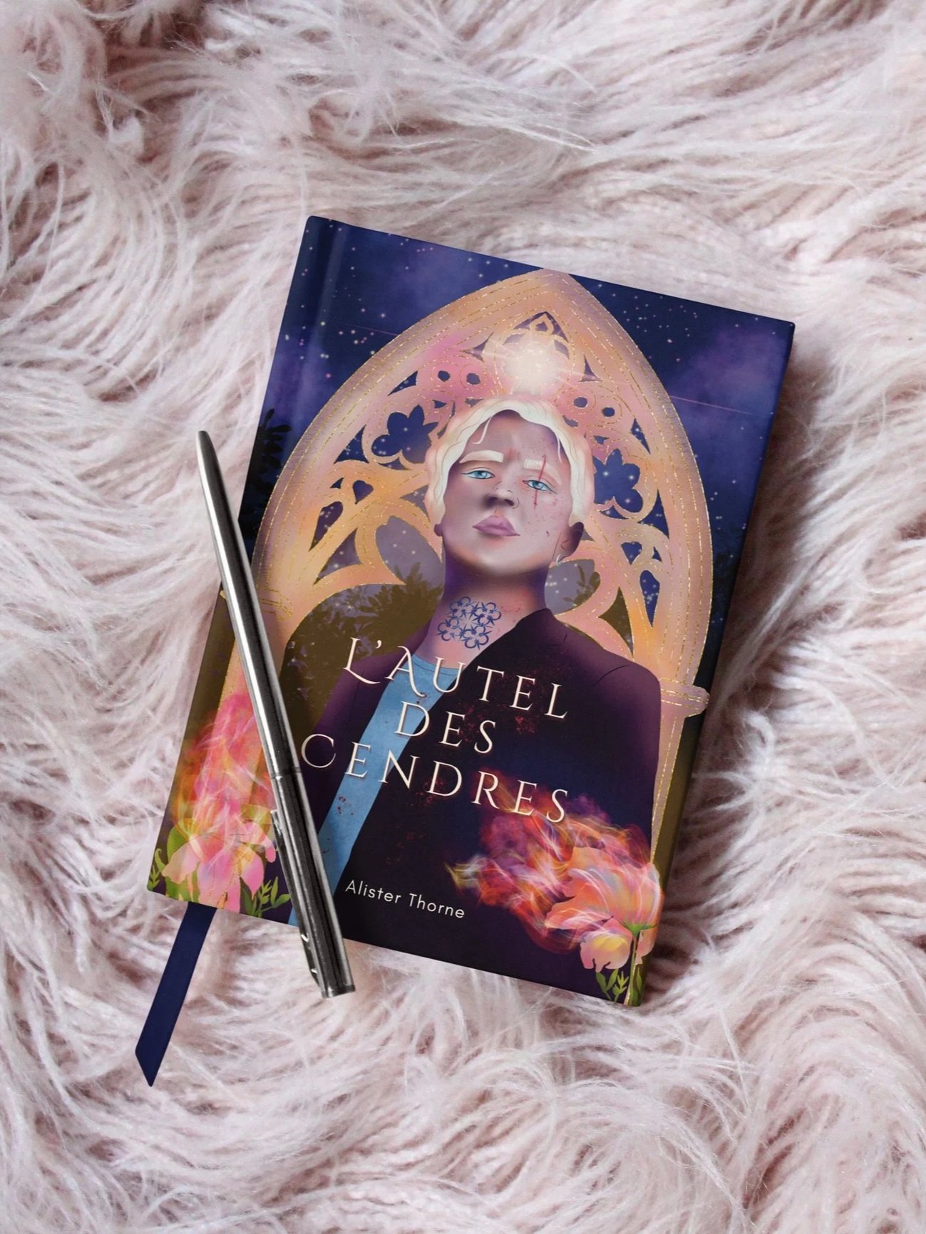 A book titled "Lautel des Cendres" by Alister Thorne, featuring a woman with short blonde hair and blue eyes, set against a mystical background with a decorative arch and pink flowers, with a silver pen resting on top, all on a pink fluffy surface.
